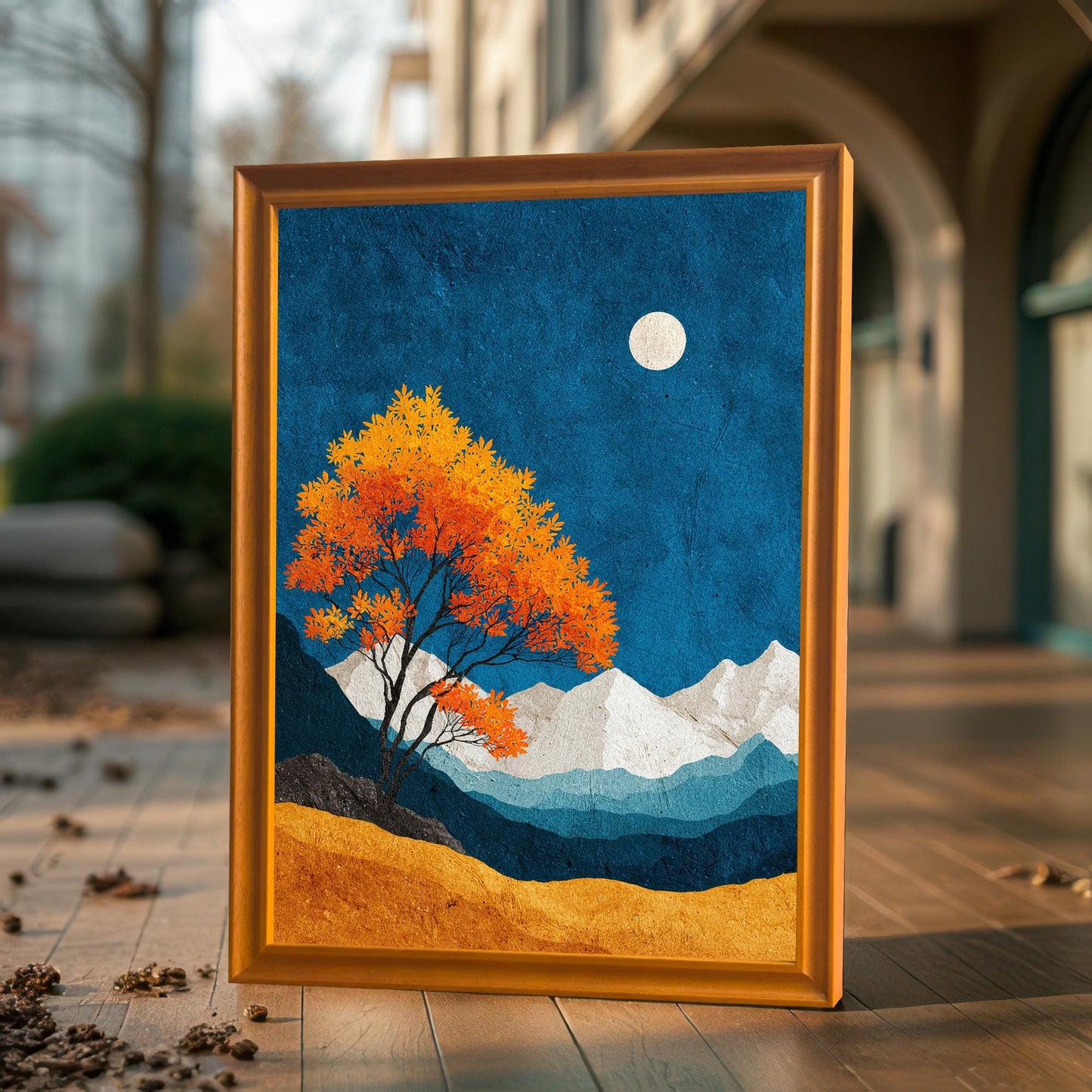A vertical art print in a natural wood frame standing on a wooden outdoor deck, reflecting warm sunlight on the textured blue and gold artwork.