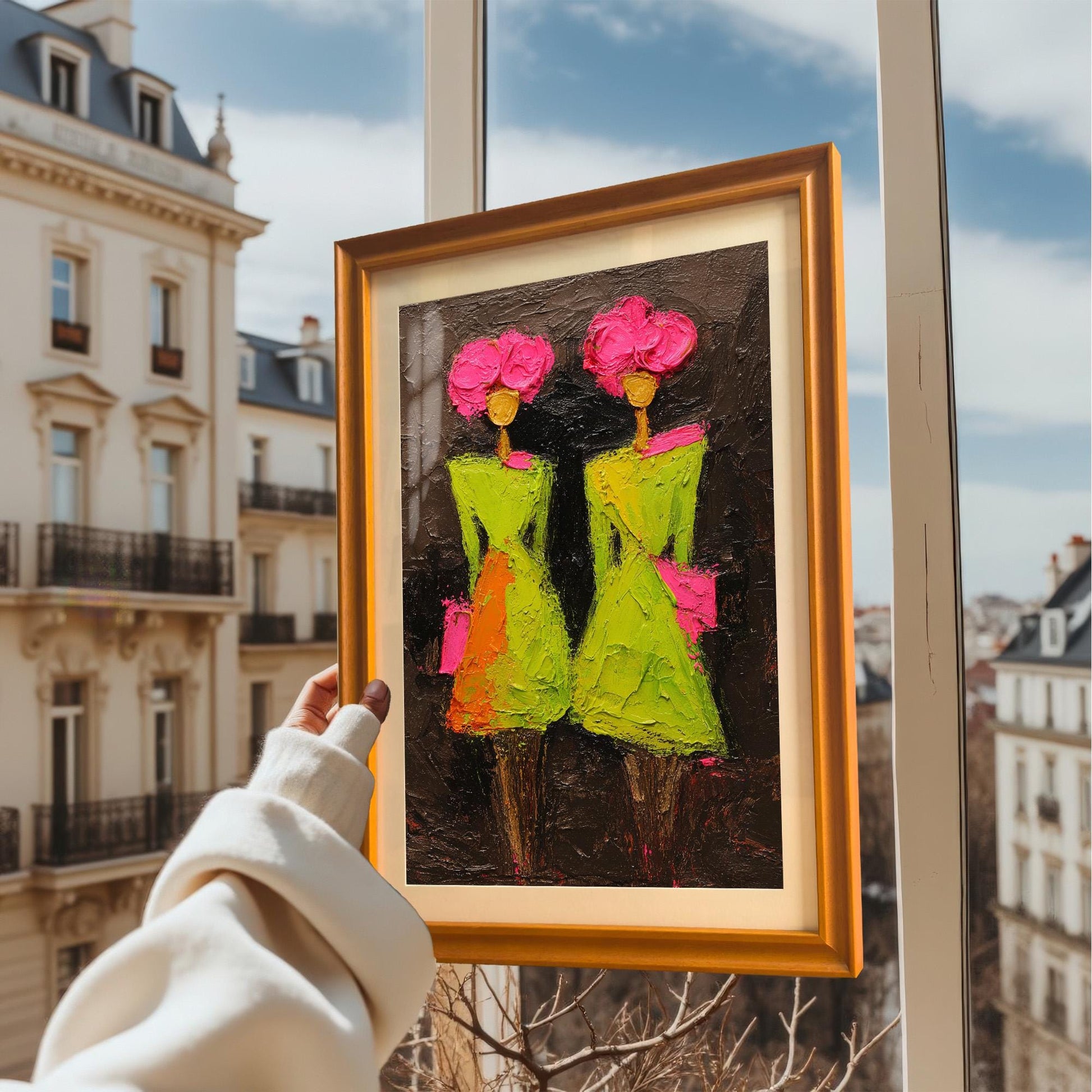A hand holding the orange-framed art print against a bright window overlooking classic European-style architecture and a blue sky.