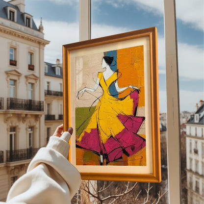 A person’s hand holding the orange-framed dancer print up against a window, showing a blurred European-style city street in the background. The natural light emphasizes the frame’s warm wood grain.