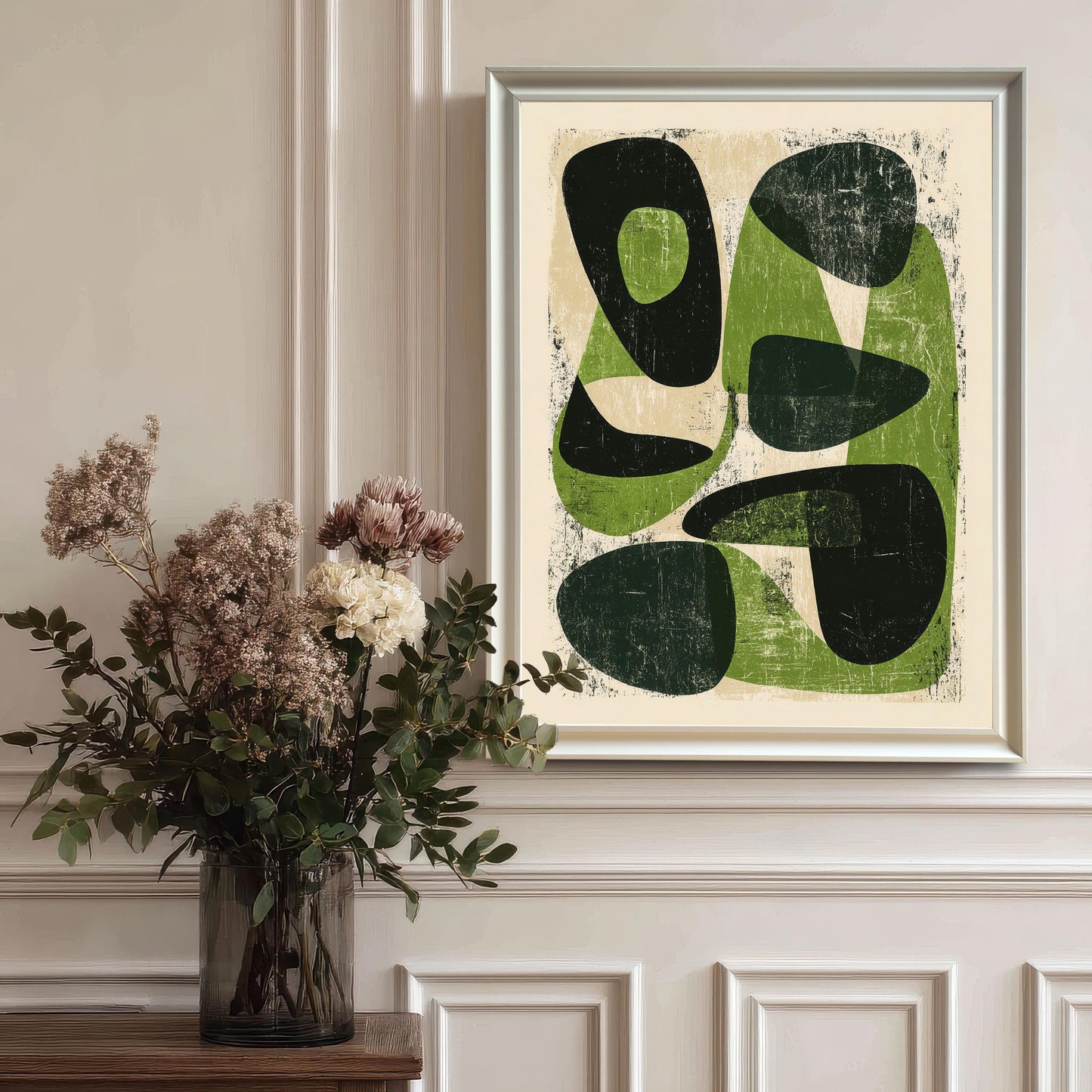The abstract green artwork in a white wood frame hanging on a classic white wainscoted wall, styled next to a glass vase with dried flowers for an elegant home decor look.