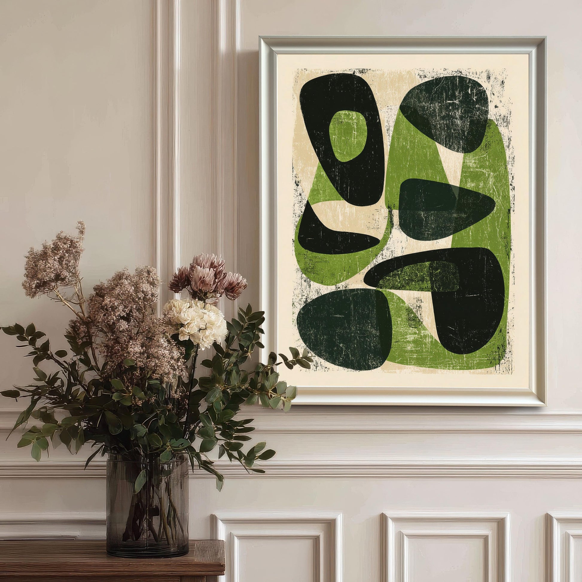 The abstract green artwork in a white wood frame hanging on a classic white wainscoted wall, styled next to a glass vase with dried flowers for an elegant home decor look.
