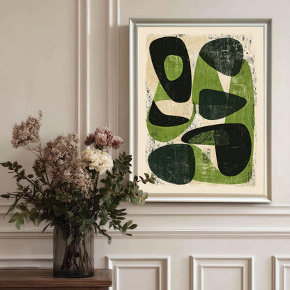 The abstract green artwork in a white wood frame hanging on a classic white wainscoted wall, styled next to a glass vase with dried flowers for an elegant home decor look.
