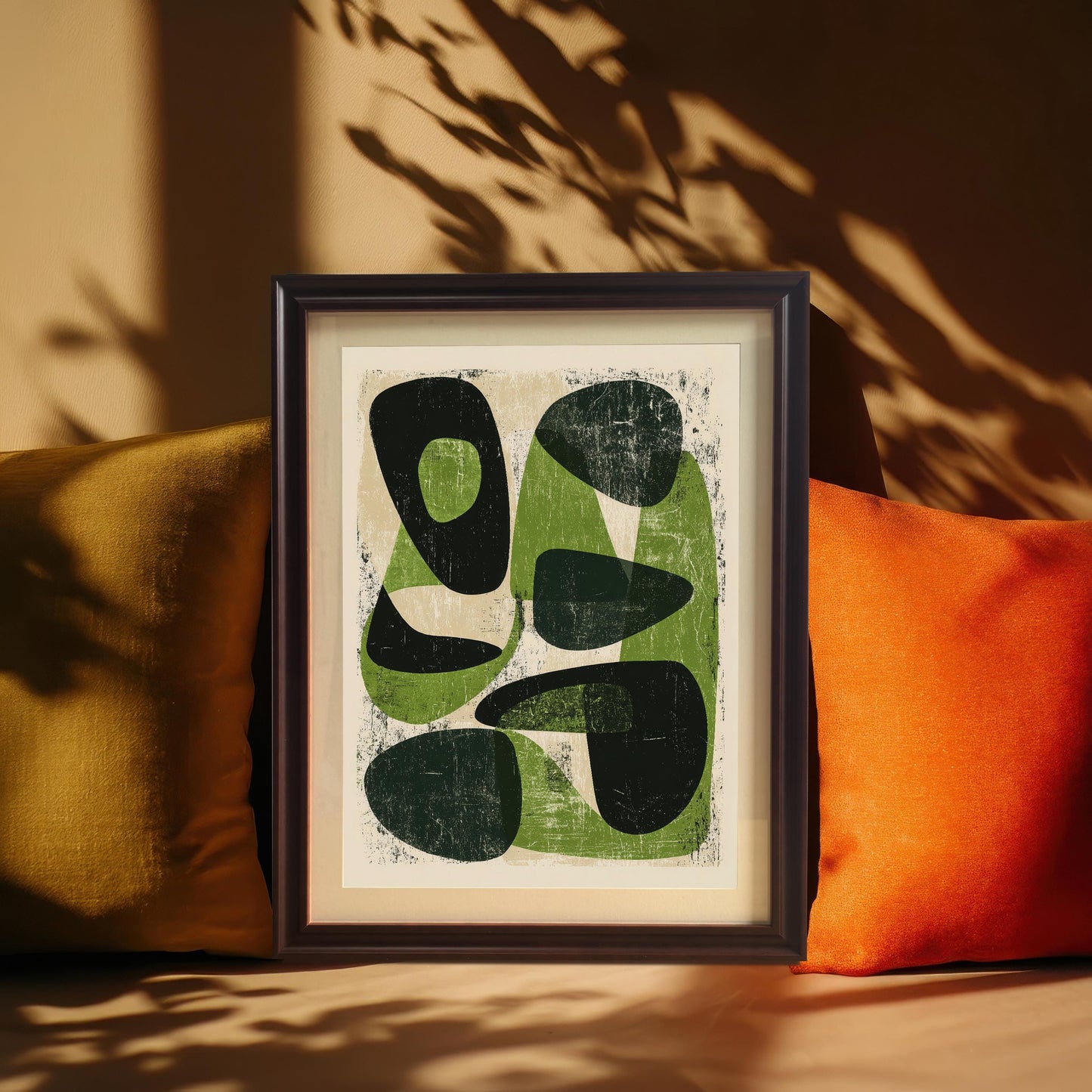 A coffee-toned wood frame containing the abstract organic print, styled between a gold and a vibrant orange velvet pillow under the play of natural leaf shadows on a tan background.
