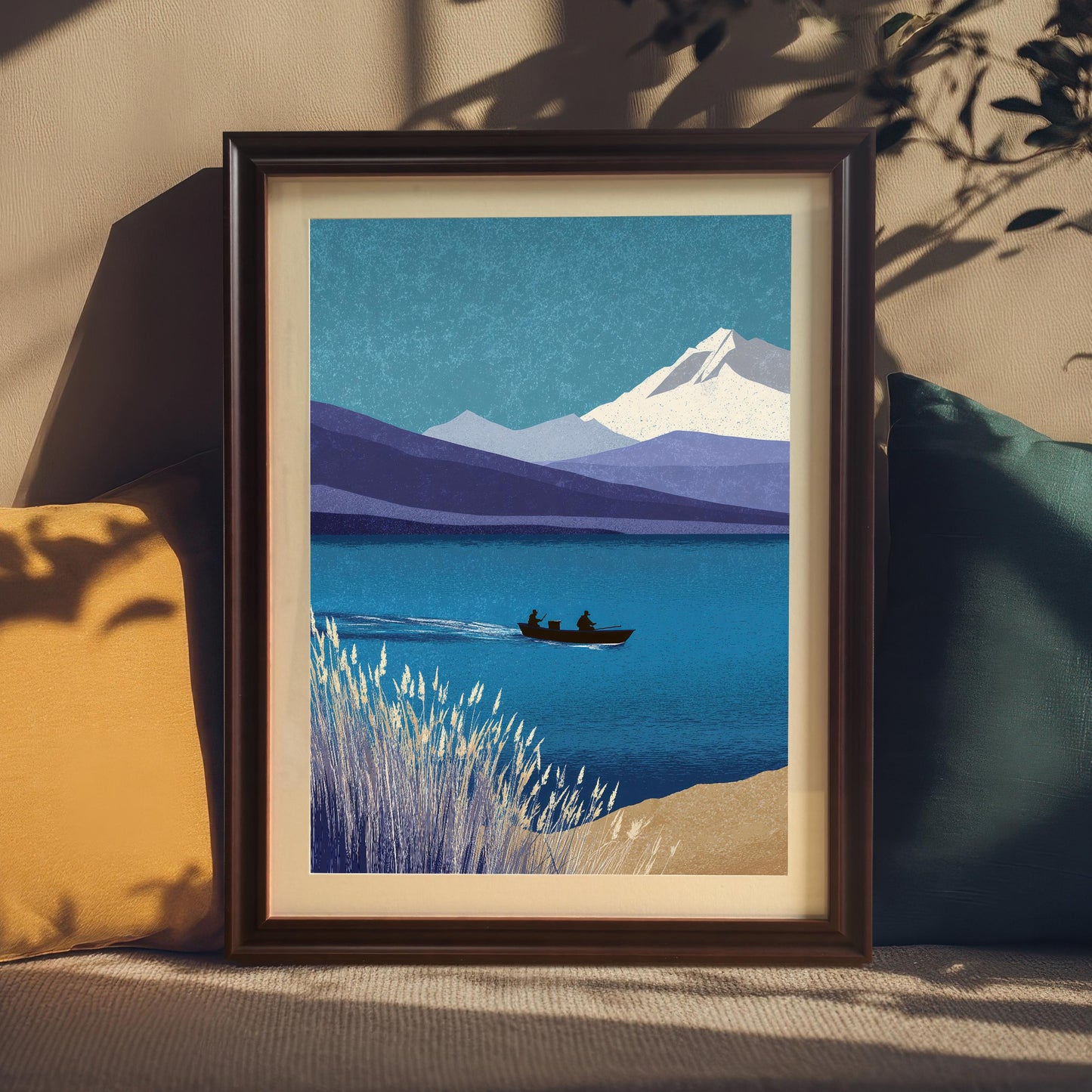 A coffee-colored wood framed art print with a cream mount, nestled between a yellow and a dark teal pillow on a textured surface. Soft sunlight and leaf shadows fall across the frame and the neutral wall behind it.
