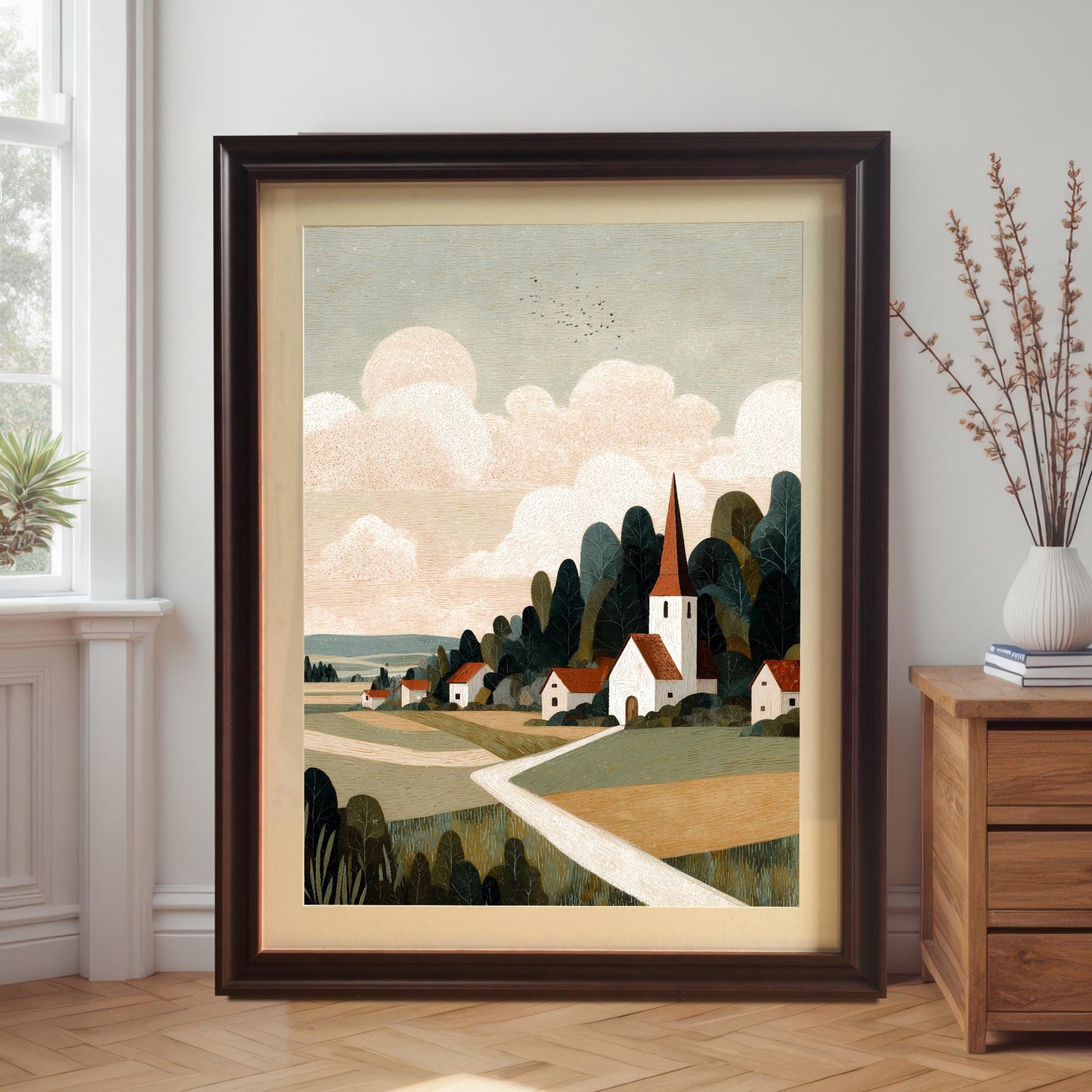 A large coffee-colored wood frame with a cream mat board surrounding the village landscape art, leaning against a white wall next to a wooden dresser and houseplant.