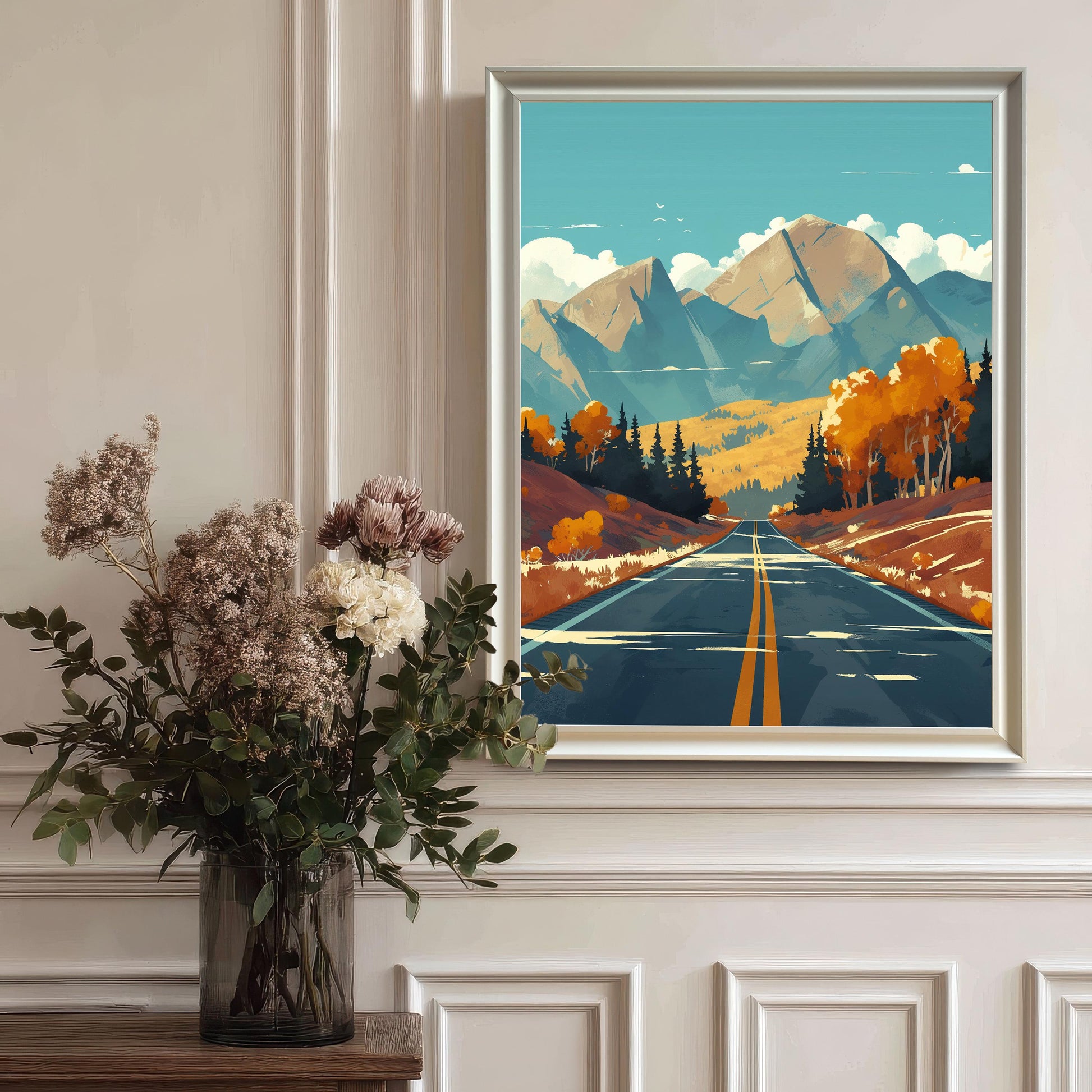 A vertical autumn mountain road art print in a sleek white wood frame, hanging on a classic white wainscoted wall behind a large vase of dried flowers.