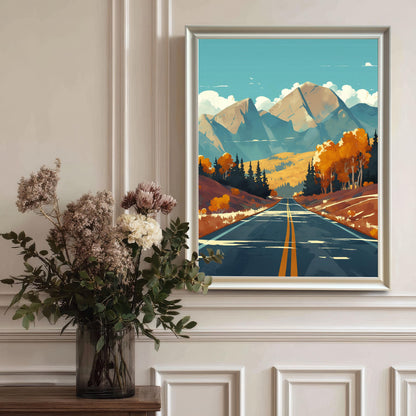 A vertical autumn mountain road art print in a sleek white wood frame, hanging on a classic white wainscoted wall behind a large vase of dried flowers.