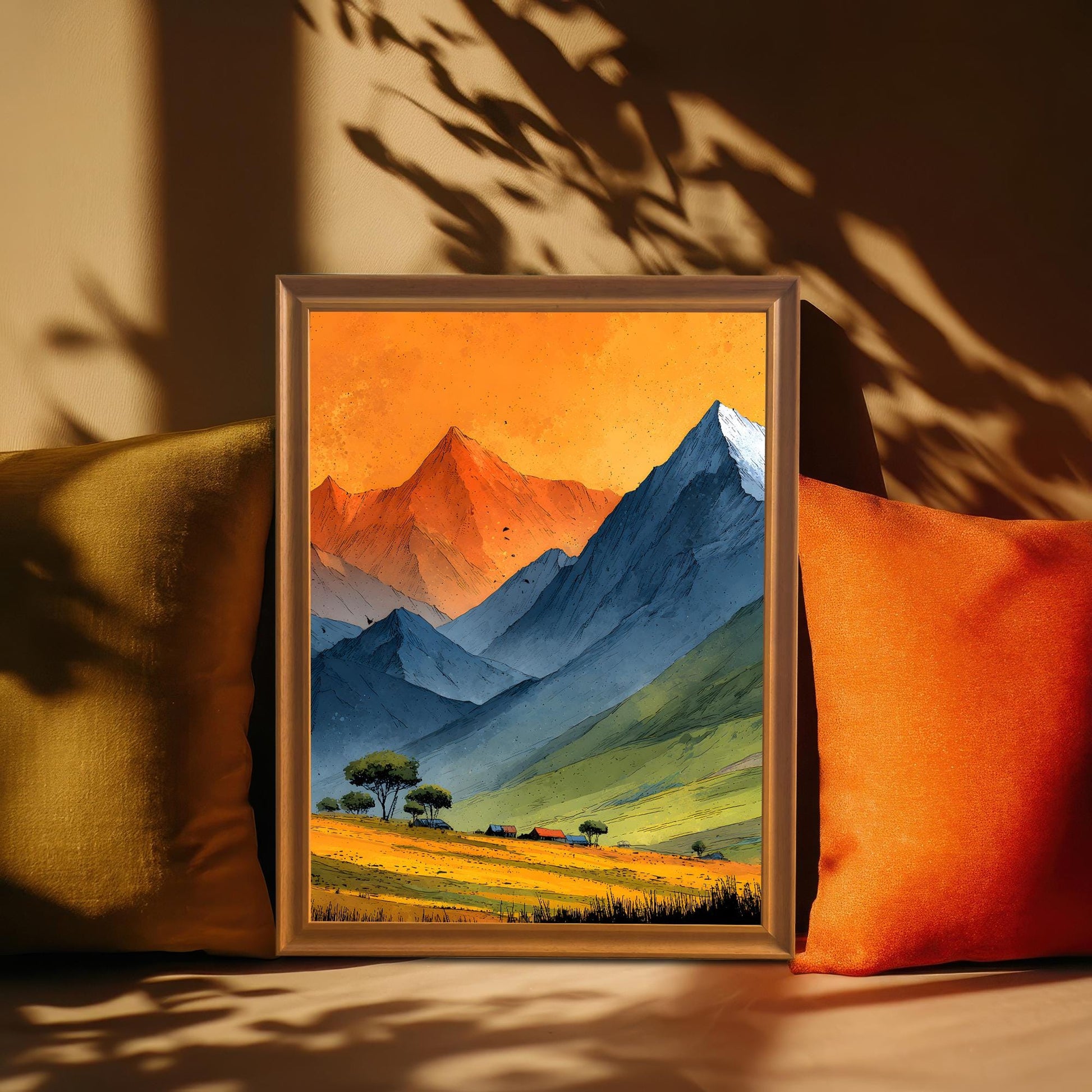 An orange-framed art print of a mountain valley tucked between yellow and orange pillows. Artistic leaf shadows are cast across the frame and the warm-toned background.