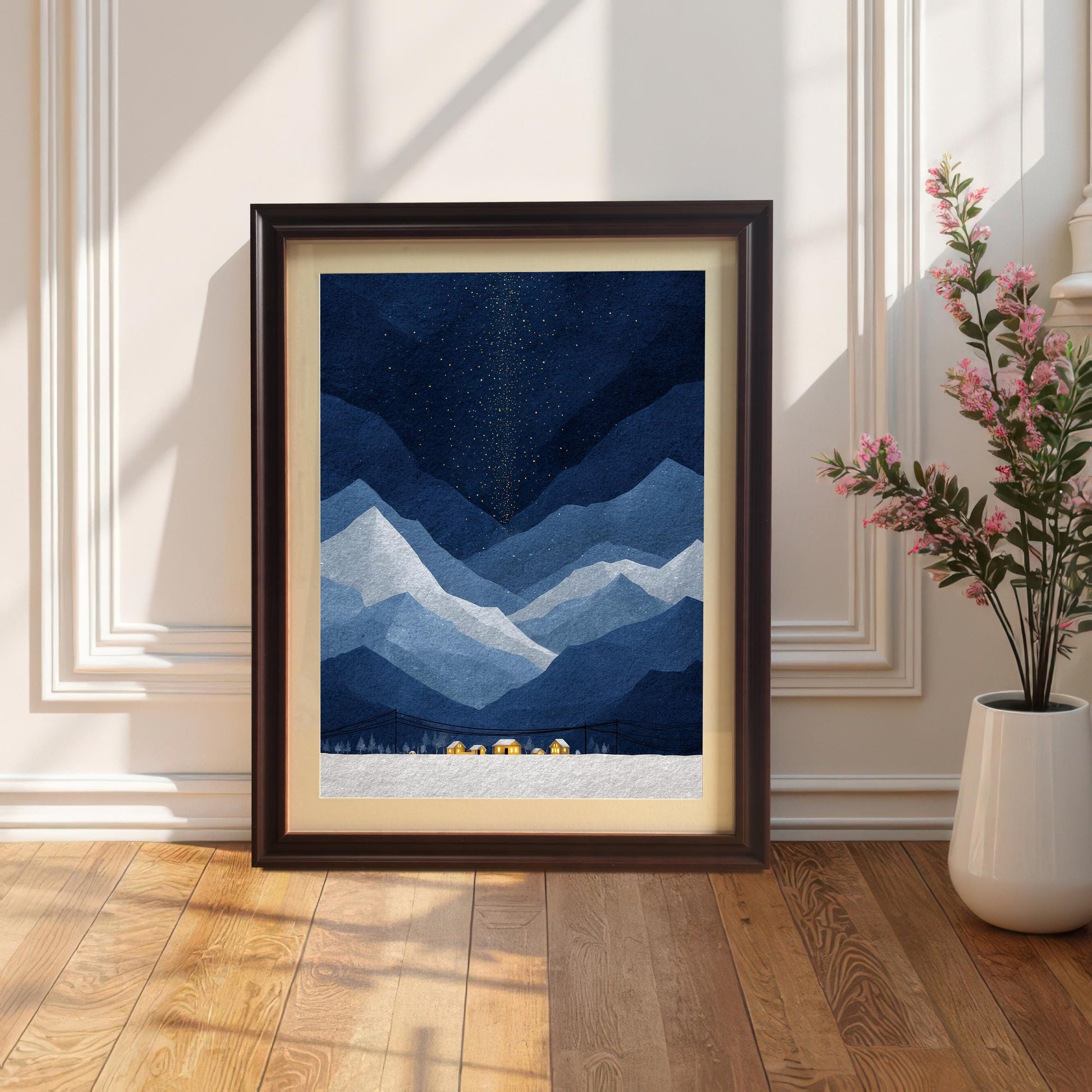 A dark coffee-wood framed mountain art print with a cream mount, leaning against a white wall with elegant molding on a polished hardwood floor next to a white vase with pink flowers.