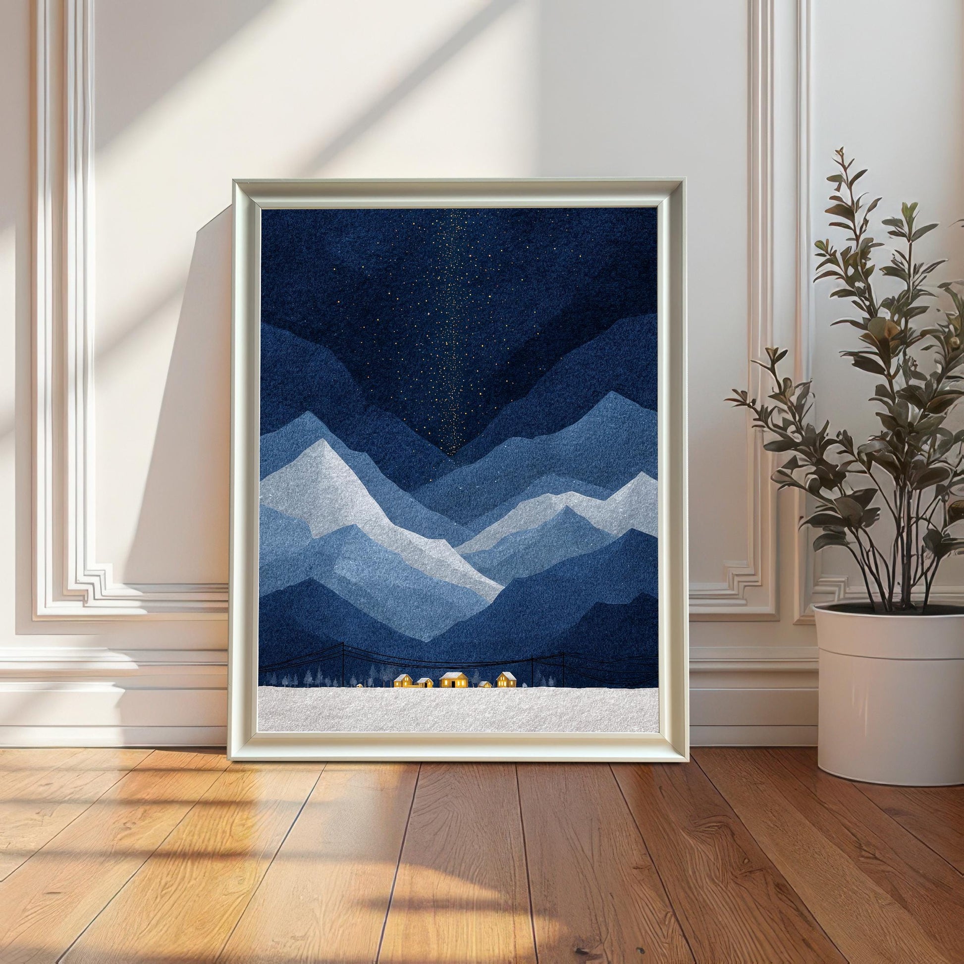 A white-framed version of the starry blue mountain landscape print, displayed without a mount, leaning against a classic white wall on a sunlit wooden floor beside a potted plant.
