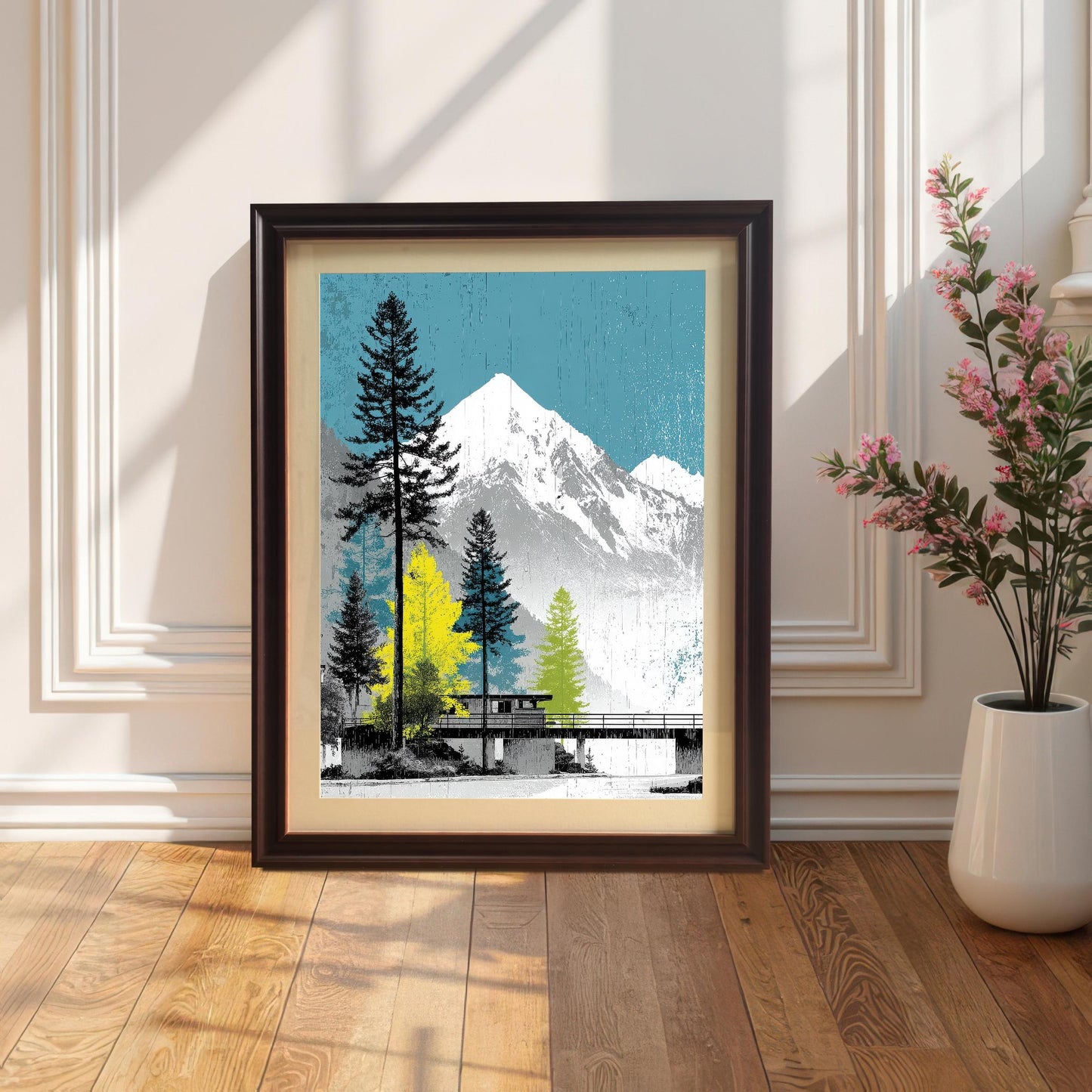 A coffee-colored dark wood frame with an ivory mount featuring a mountain landscape art print, leaning against a white paneled wall next to a vase of pink flowers.