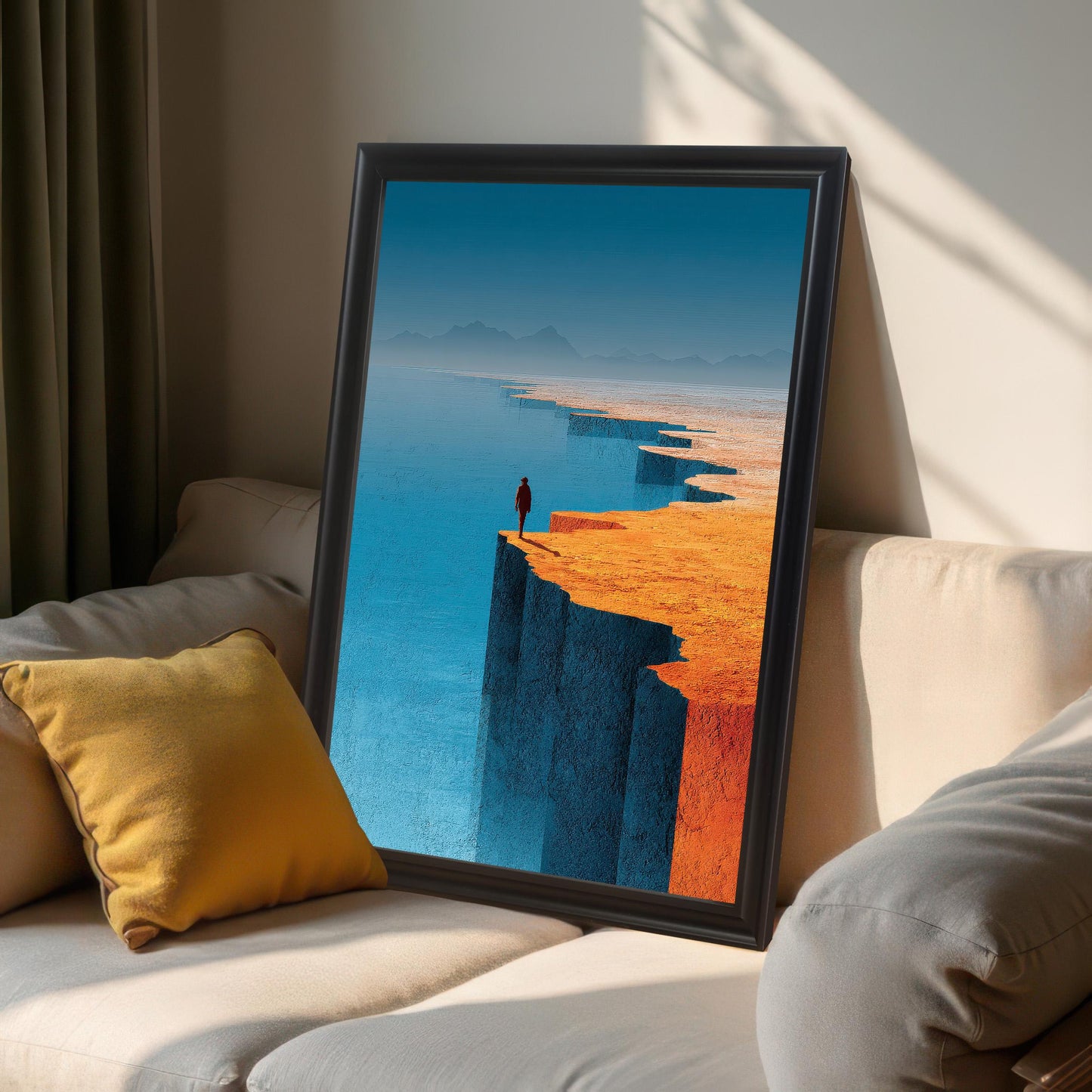 A large, black-framed art print of a person standing on a cliff edge leaning against a cream-colored sofa in a sunlit room. A yellow accent pillow sits on the couch next to the frame.