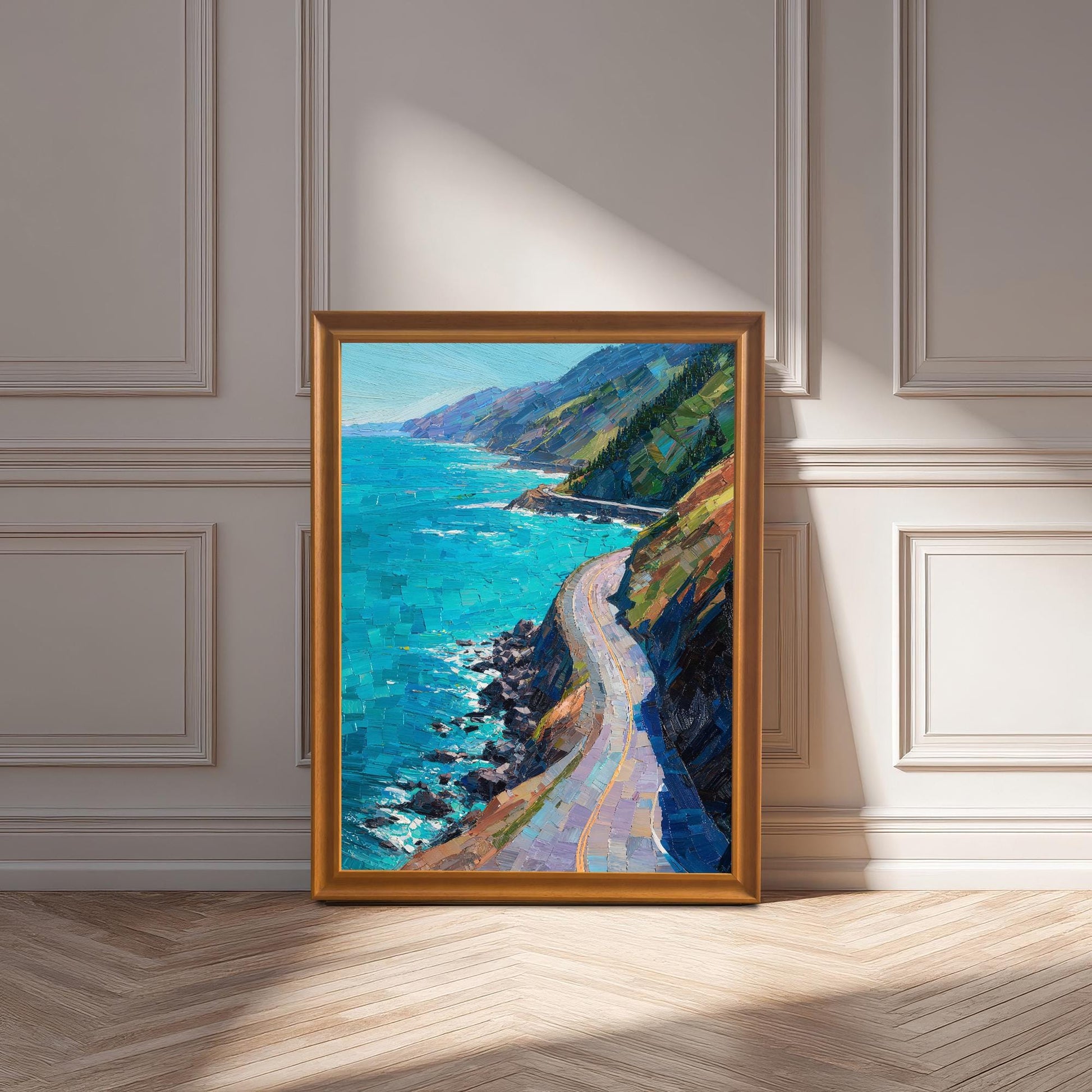 An Orange wood framed vertical landscape print of a winding coastal road and turquoise sea, leaning against an elegant white wainscoted wall.