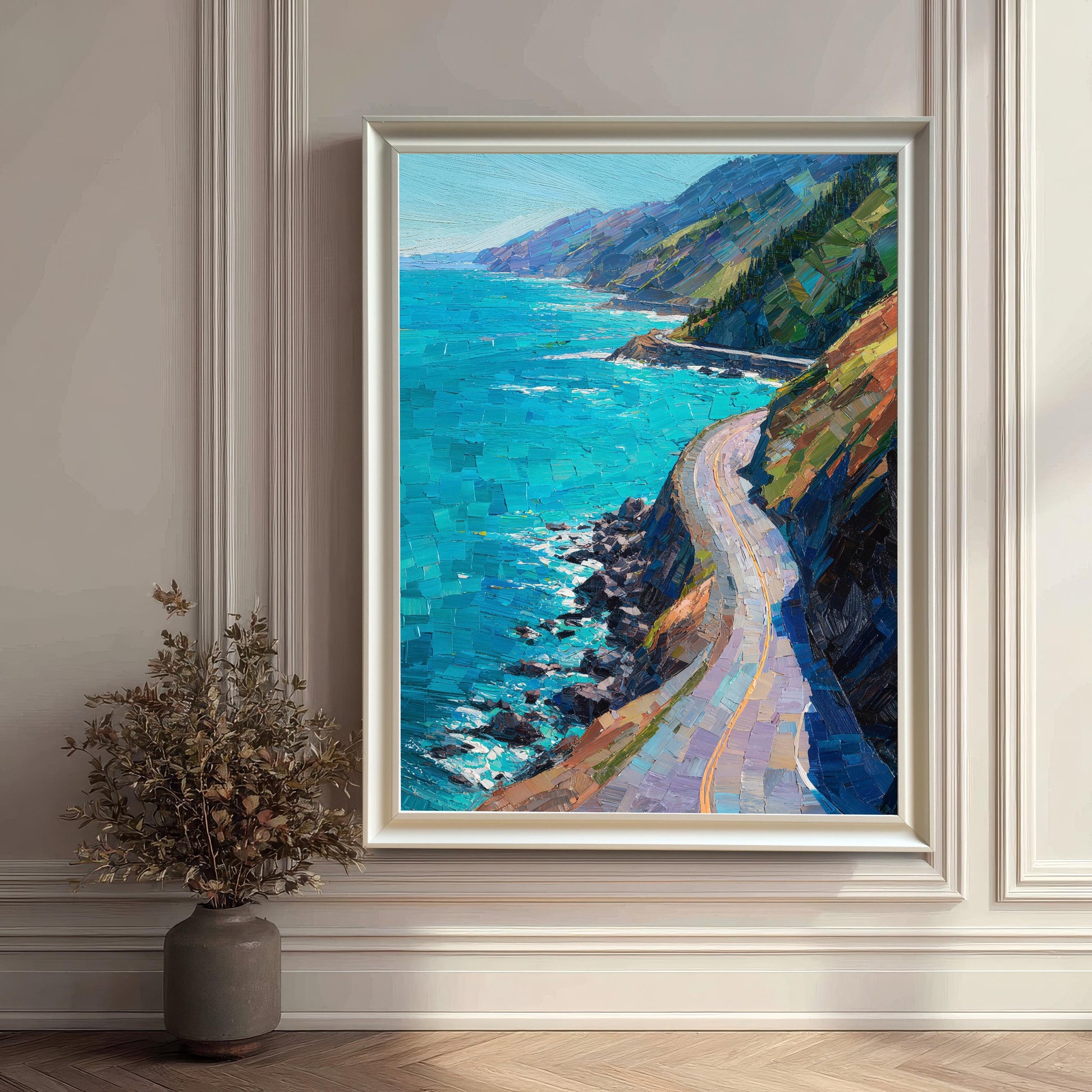 A white wood framed vertical art piece showcasing a vibrant blue ocean and coastal highway, mounted on a white wainscoted wall next to a dark gray vase with decorative dried branches.