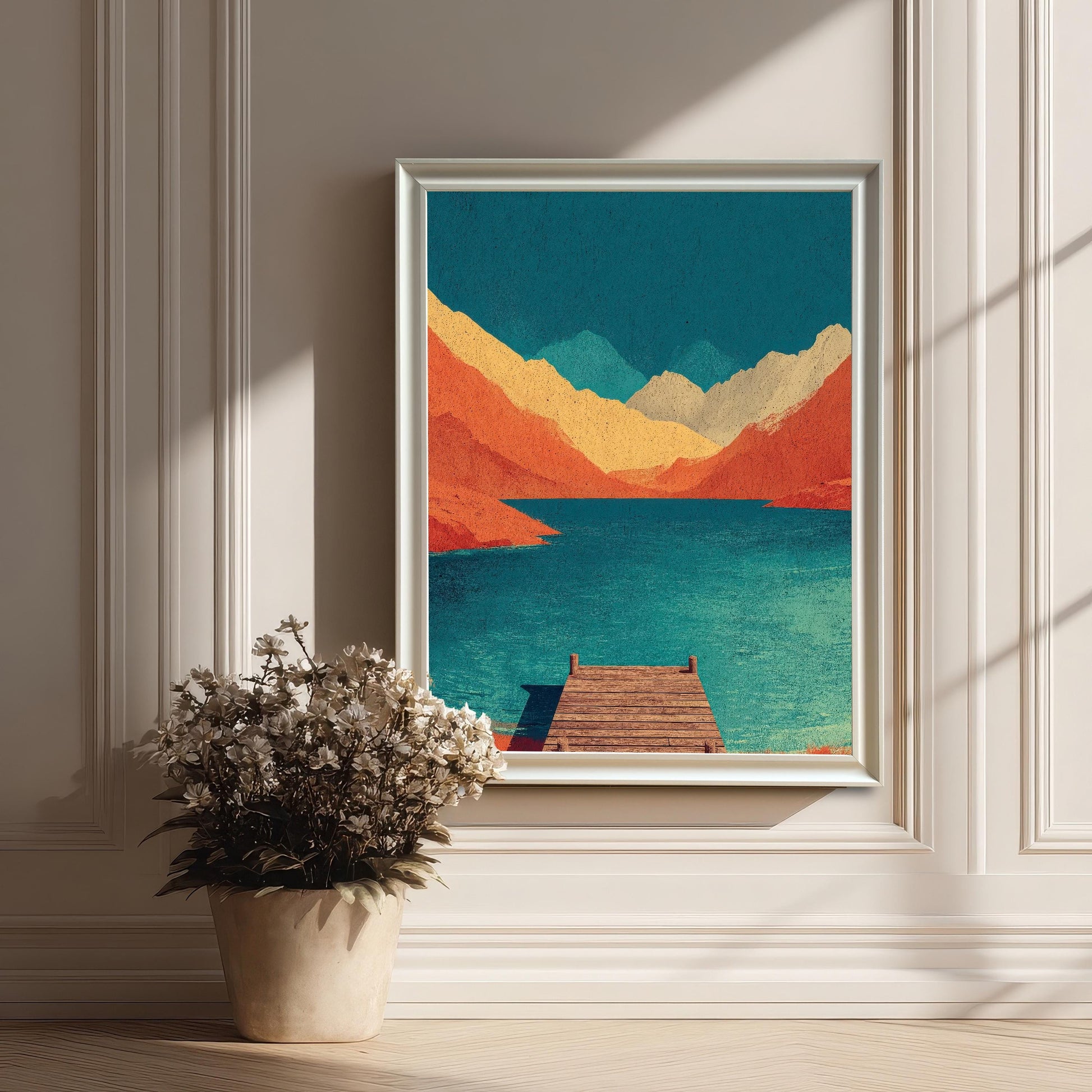 A white framed abstract mountain lake art print sitting on a ledge of a classic white wainscoted wall next to a pot of white flowers.