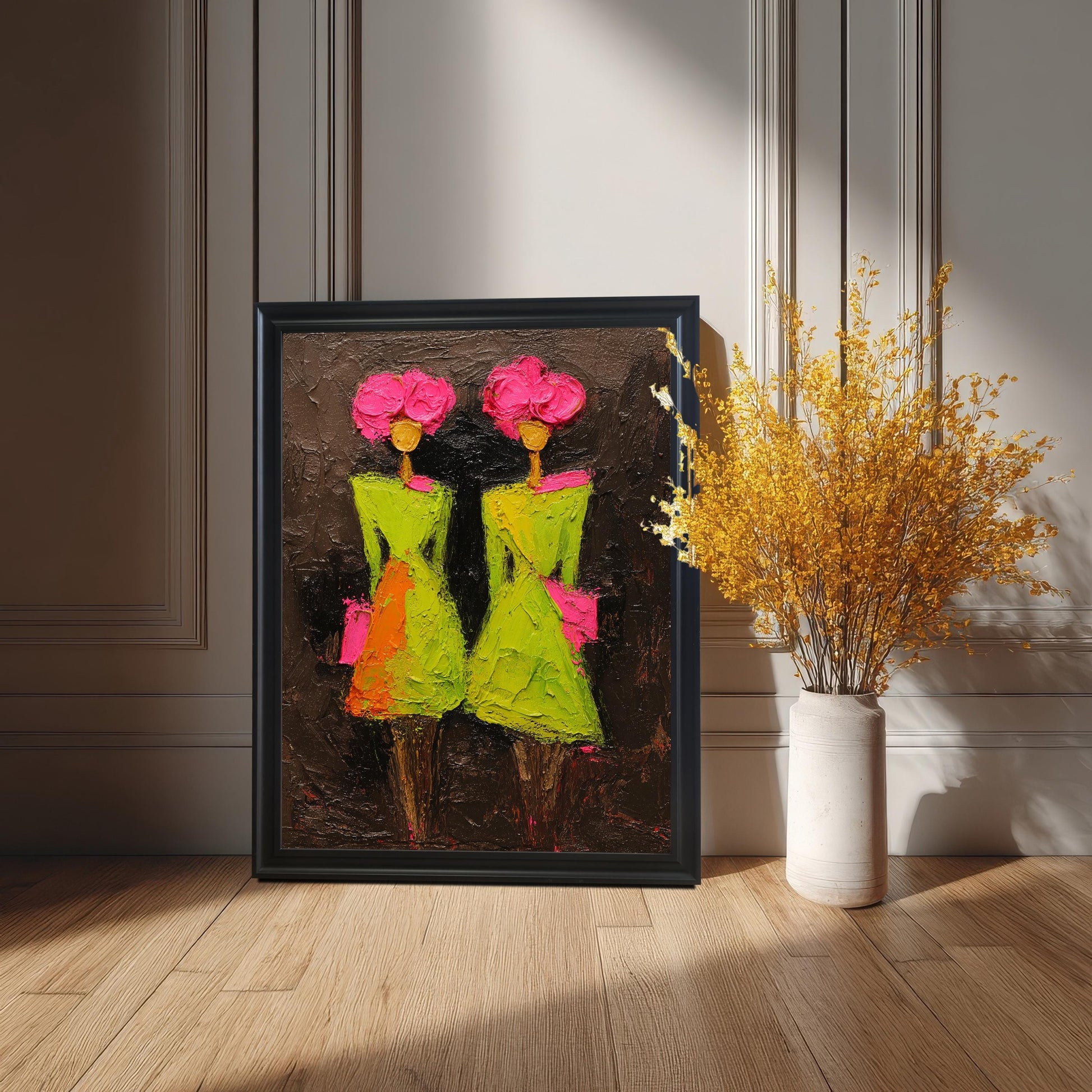 A black-framed art print of two stylized figures in green and pink leaning against a light-colored paneled wall on a light wood floor, placed next to a tall white vase filled with bright yellow dried flowers.