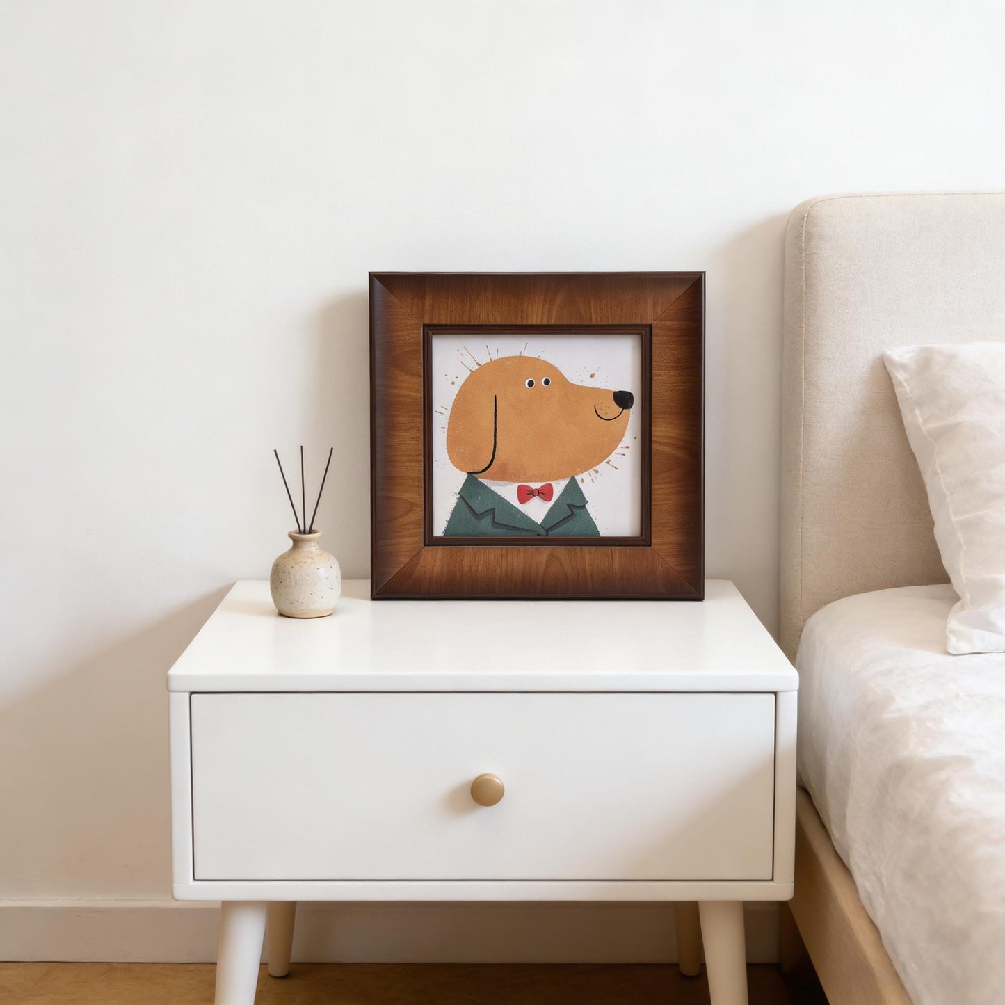 A square framed whimsical dog illustration sitting on a white bedside table next to a small ceramic vase with diffuser sticks.