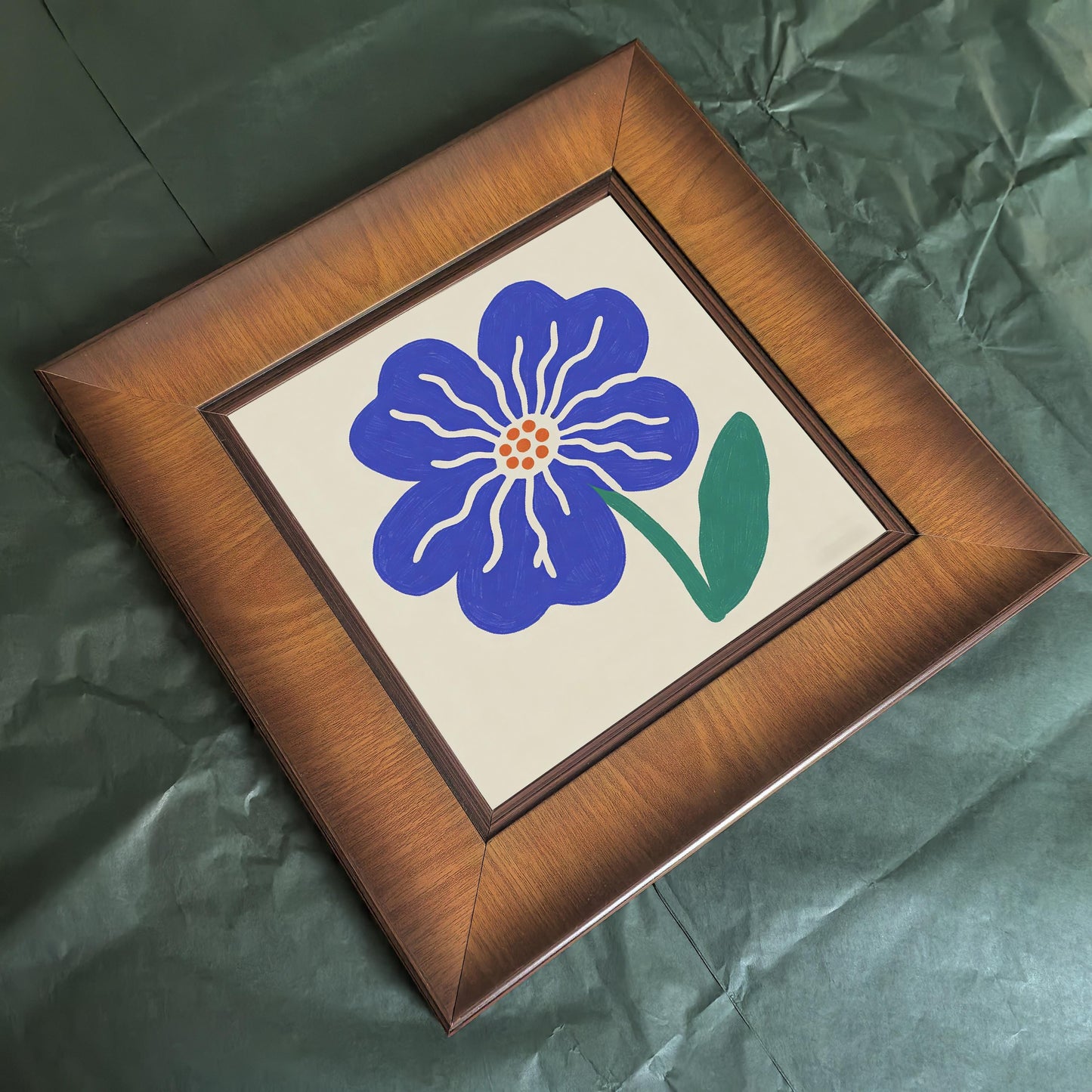 Top-down view of a square wooden frame with a blue flower print lying on a dark green textured surface, highlighting the rich wood finish.