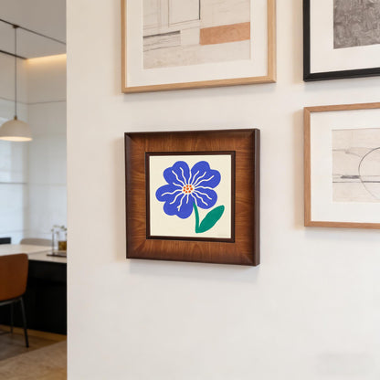 Square dark wood-framed flower print hanging on a white gallery wall alongside other modern minimalist art pieces in a contemporary home.