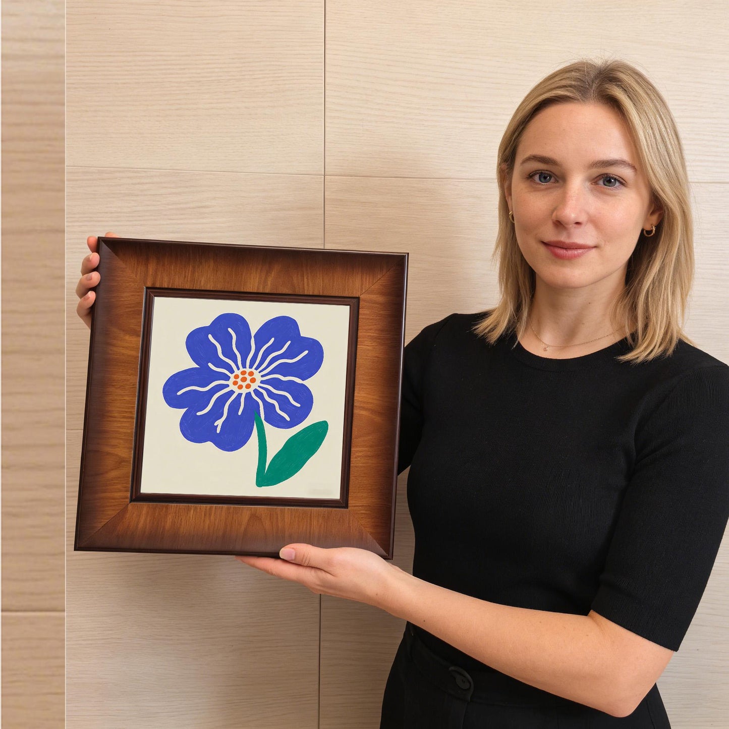 A woman holding the square blue flower art print to show scale; the dark wood frame complements her black top and the neutral background.