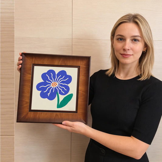 A woman holding the square blue flower art print to show scale; the dark wood frame complements her black top and the neutral background.