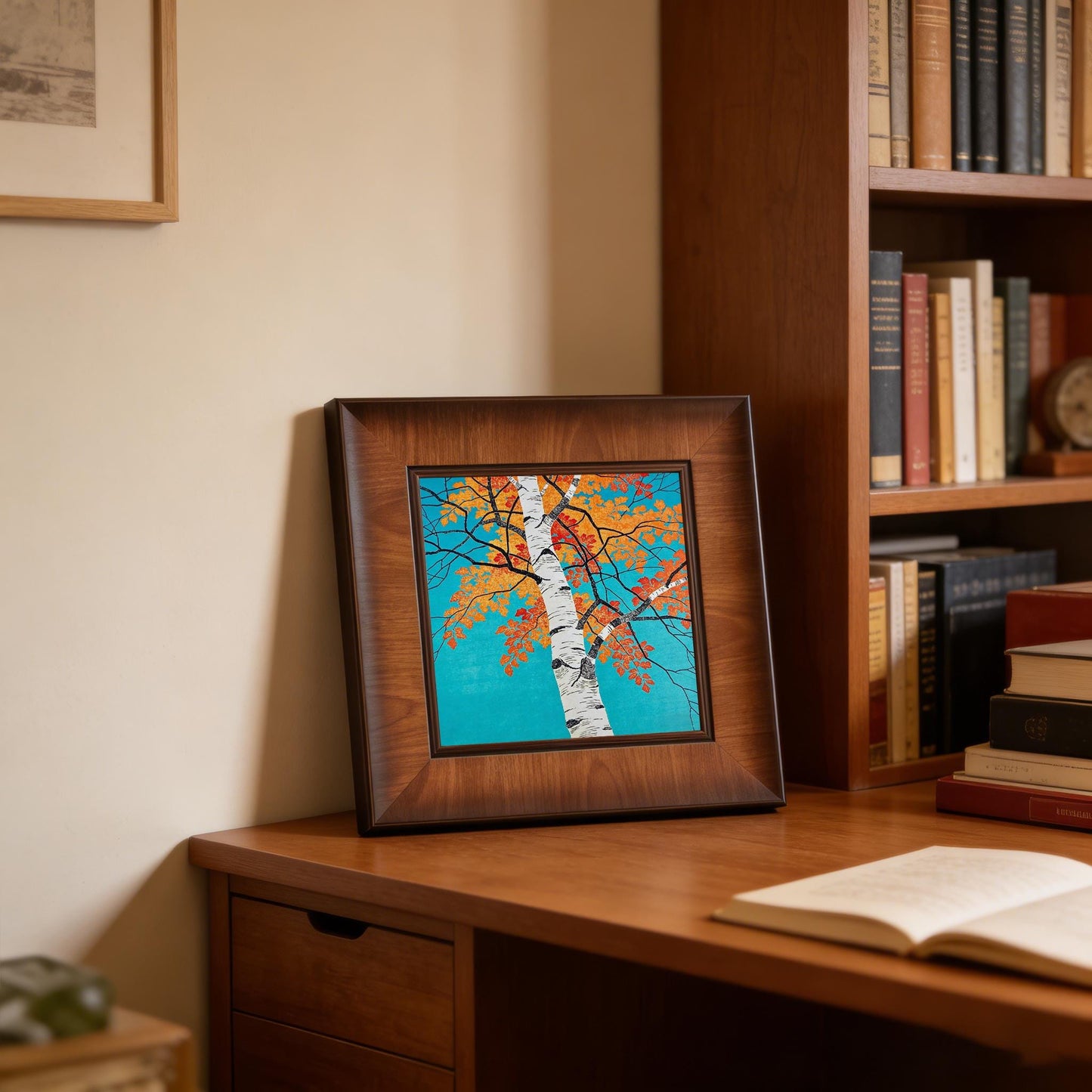 The autumn birch tree framed artwork displayed on a wooden desk next to a bookshelf, creating a cozy and scholarly home office atmosphere.v