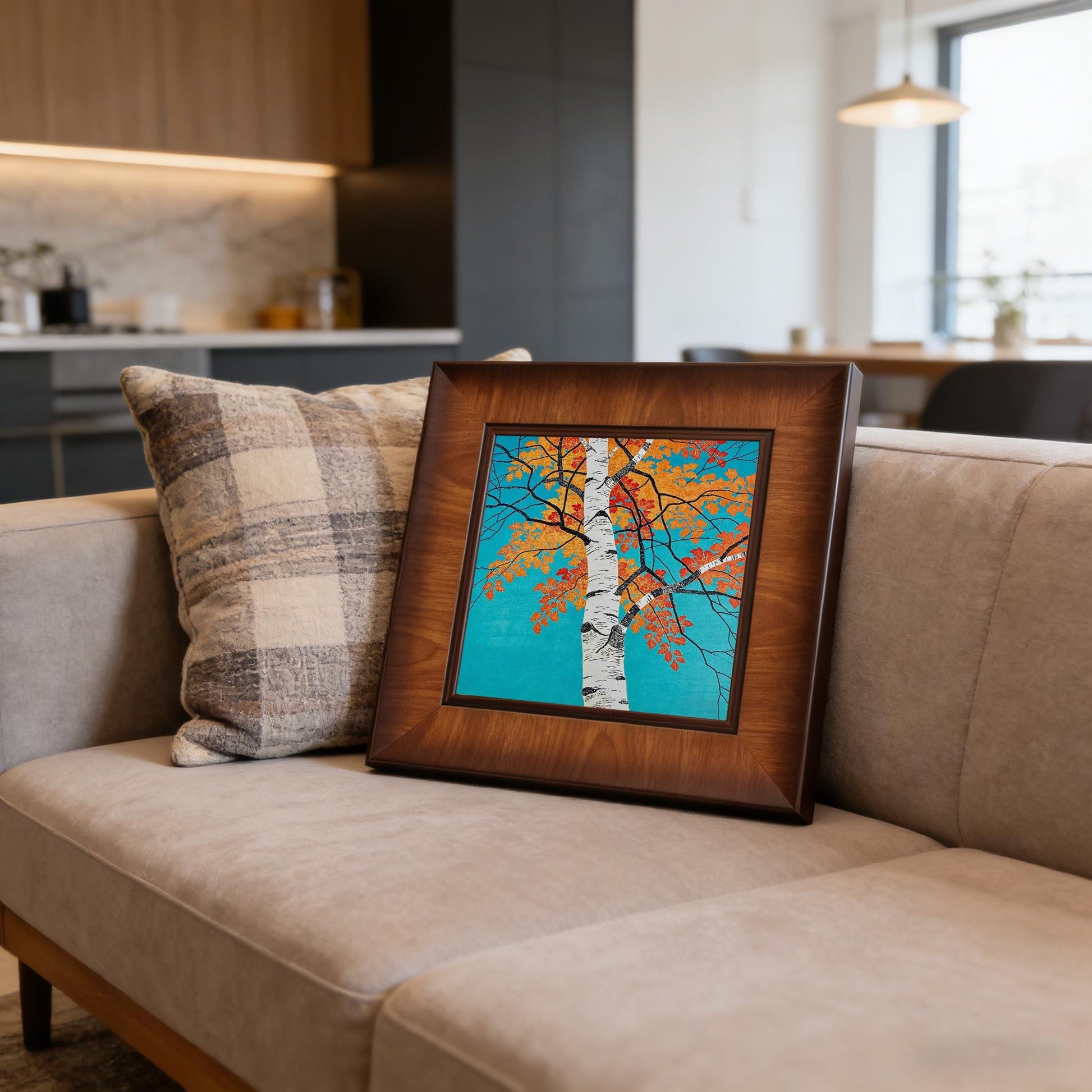 The framed autumn tree artwork resting on a modern grey sofa next to a plaid pillow, showcasing how the piece complements contemporary living room decor.