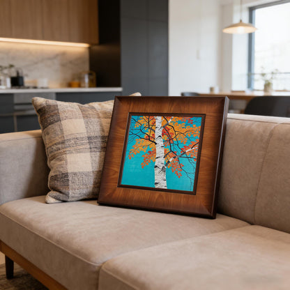 The framed autumn tree artwork resting on a modern grey sofa next to a plaid pillow, showcasing how the piece complements contemporary living room decor.