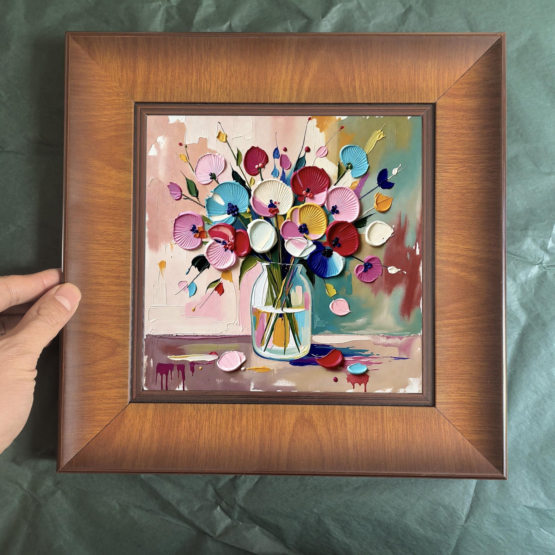 A hand holding a square wood-framed floral print against a dark green textured background, showcasing the 3D impasto details of the colorful flower vase.