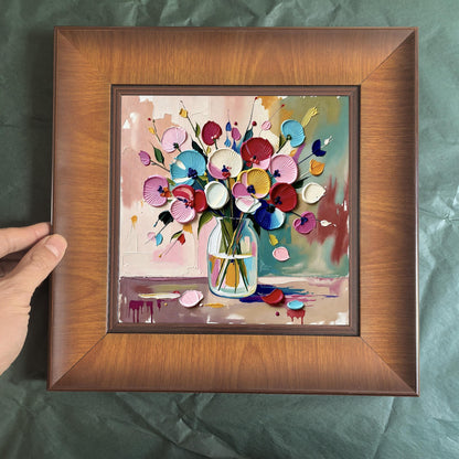 A hand holding a square wood-framed floral print against a dark green textured background, showcasing the 3D impasto details of the colorful flower vase.