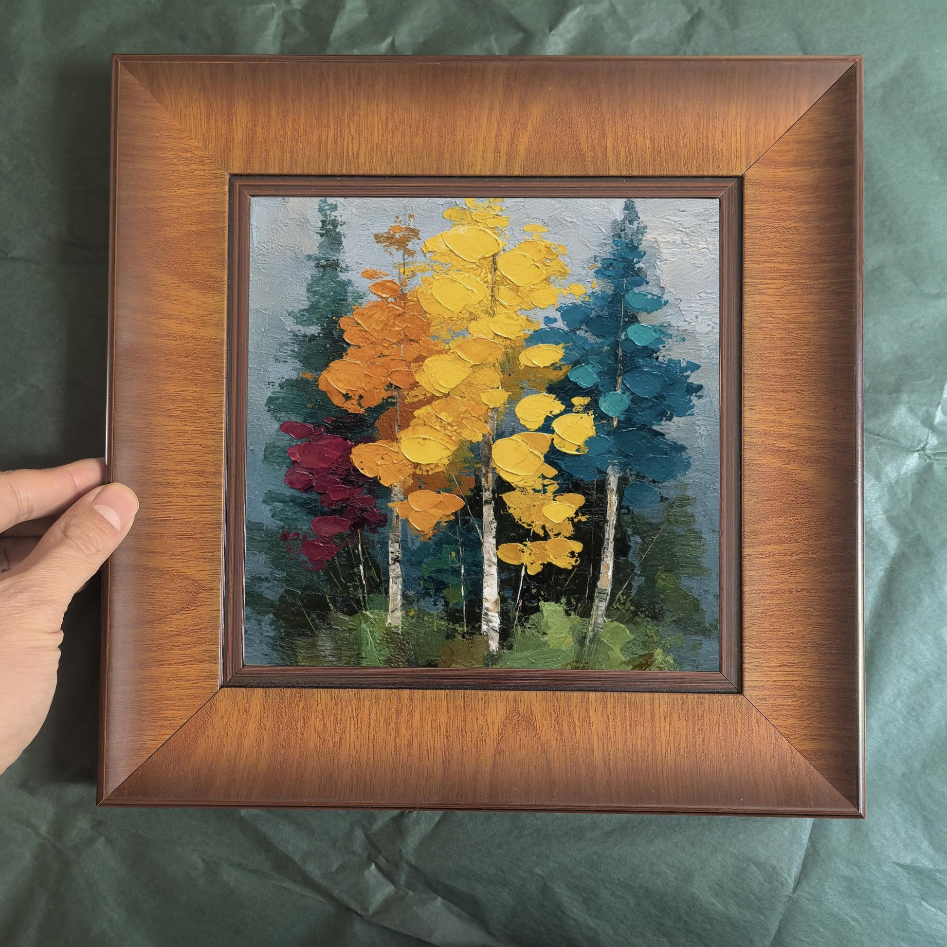 Close-up of a hand holding a square wood-framed painting of vibrant yellow and blue birch trees against a green background.