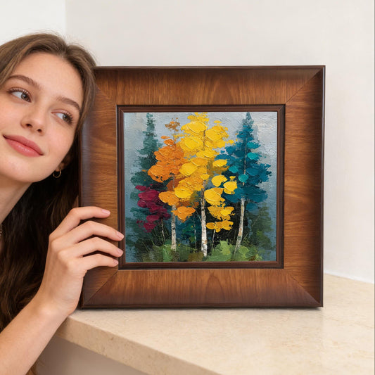 A woman smiling and posing next to a square, dark wood-framed impasto painting of a colorful autumn forest.
