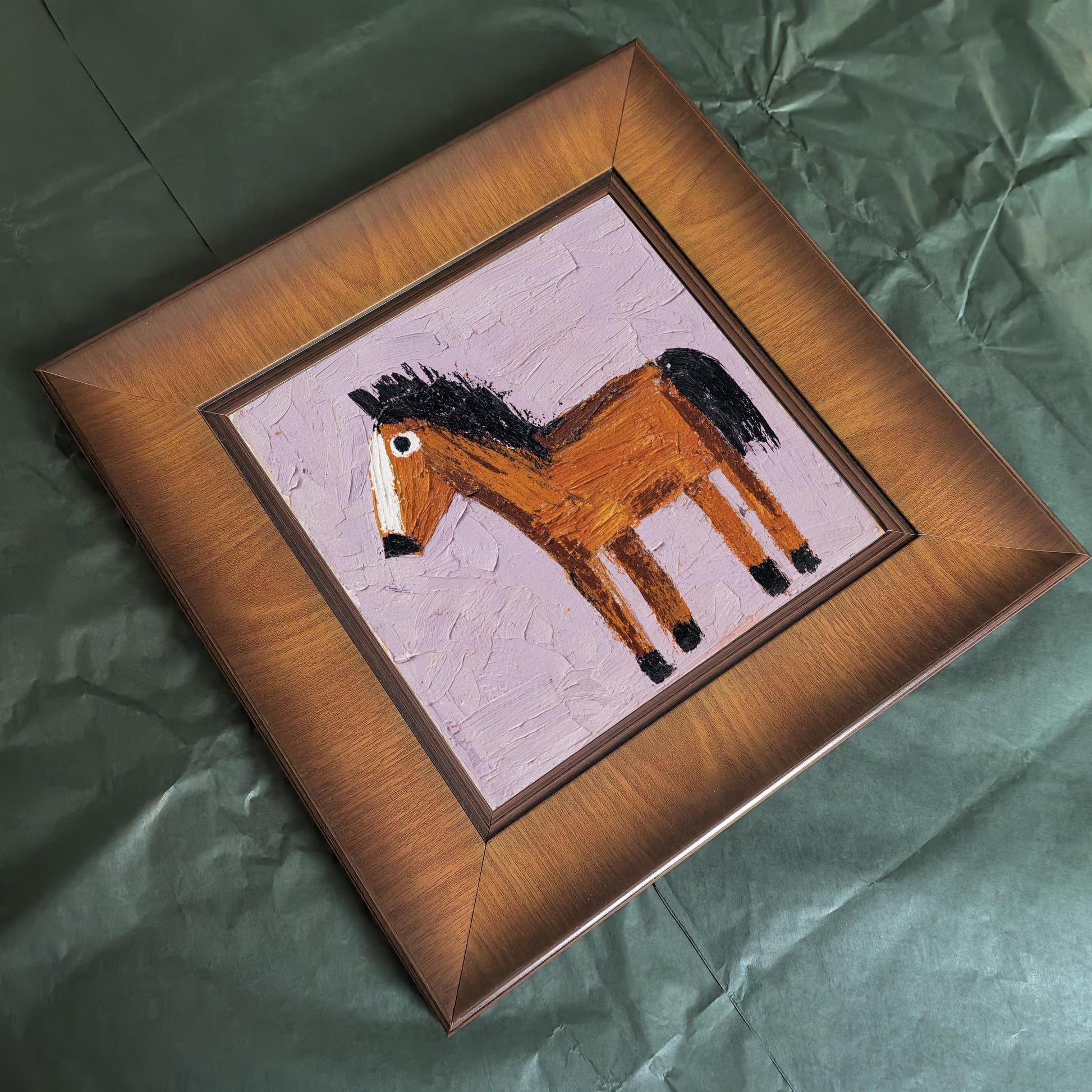 High-angle shot of the square wood-framed horse painting lying flat on a textured dark green background, highlighting the beveled frame edges and the thick impasto texture of the pink background.