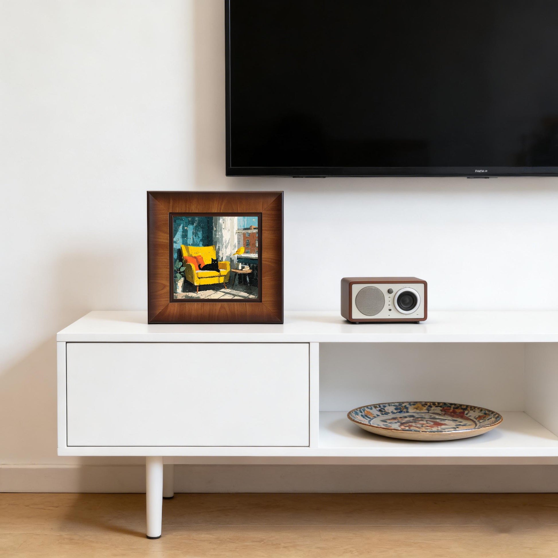 Framed whimsical cat art sitting on a white TV stand next to a vintage-style radio in a bright living room.