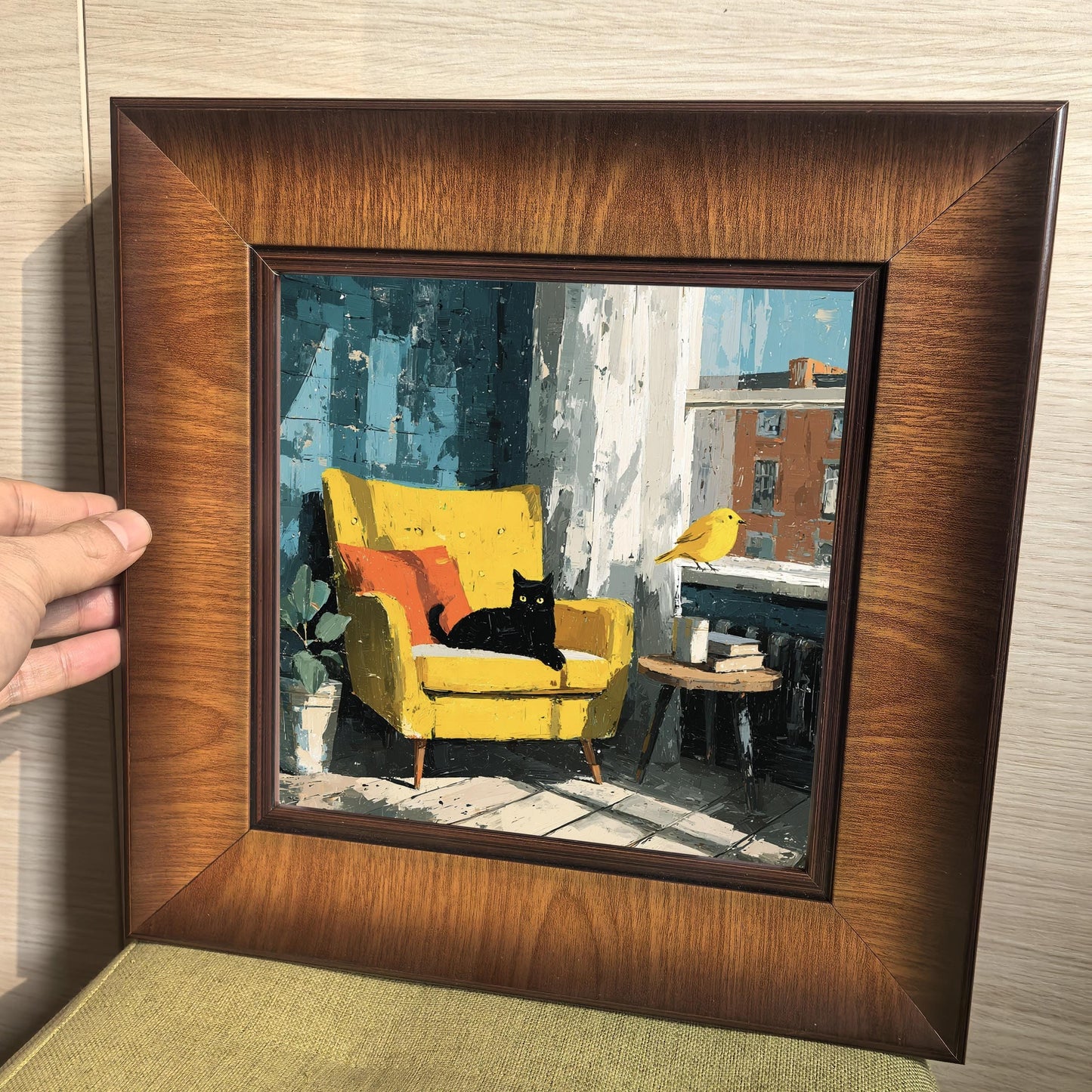 Hand holding a square walnut-finish wood frame to show the scale and texture of the cat and yellow chair painting.