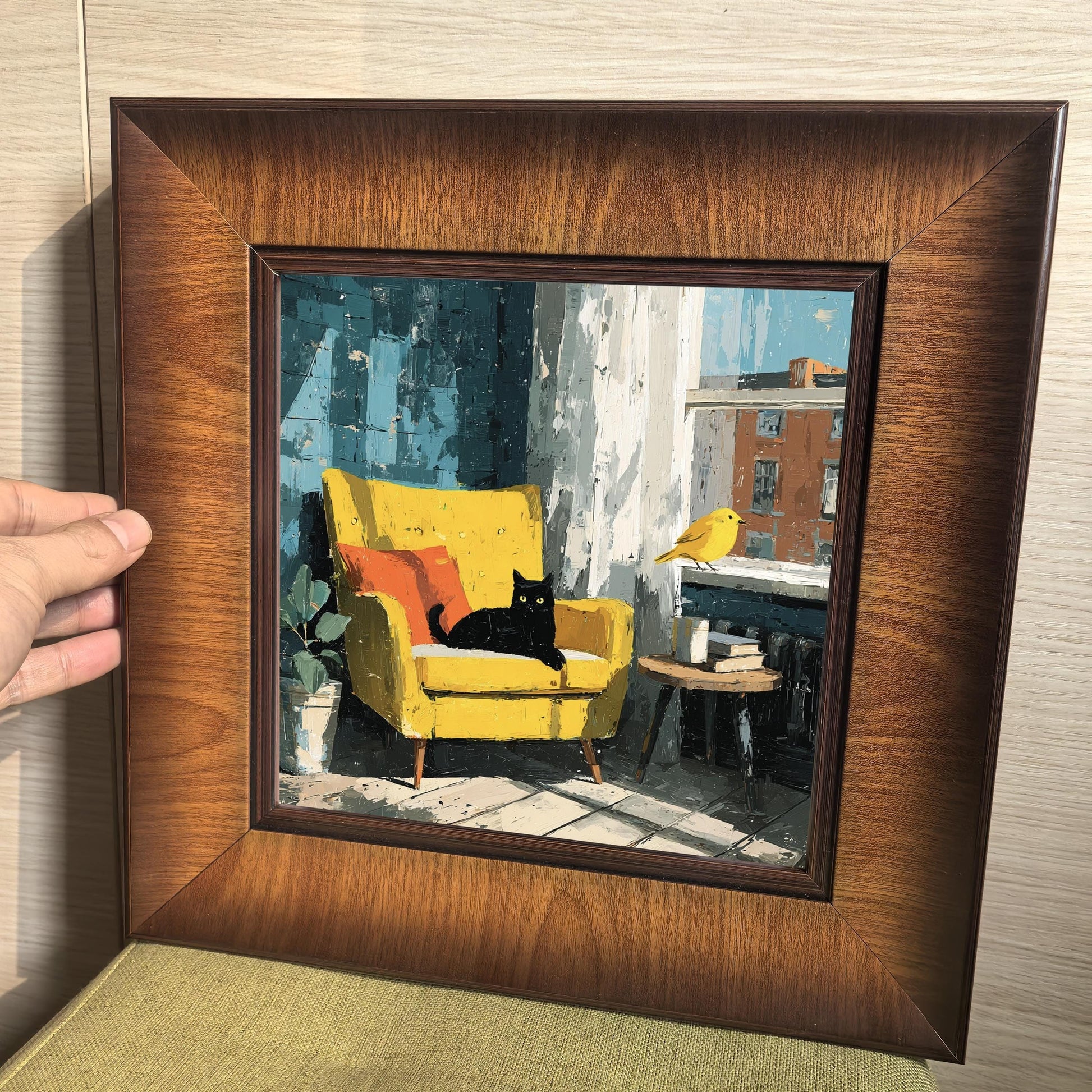Hand holding a square walnut-finish wood frame to show the scale and texture of the cat and yellow chair painting.