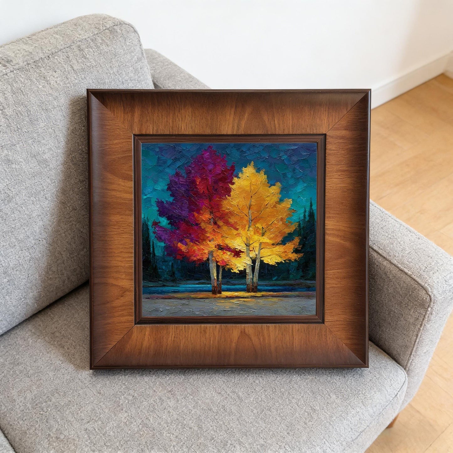 Square dark wood-framed autumn landscape resting on a grey fabric couch, demonstrating how the art looks in a contemporary living room.