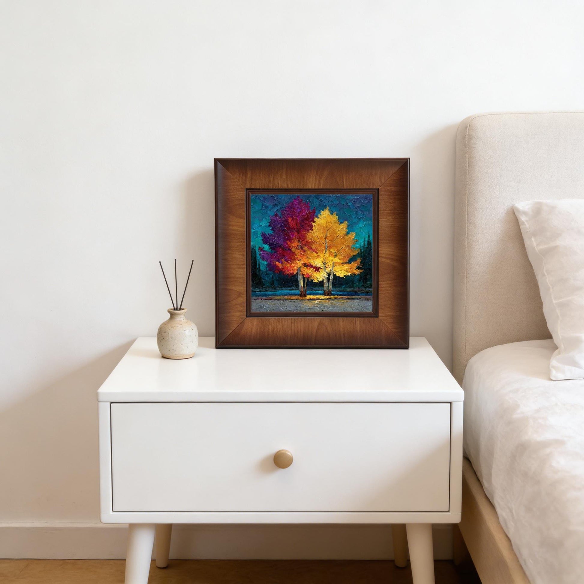 Colorful square wood-framed art leaning on a white nightstand next to a bed, styled with a small ceramic diffuser for a cozy home feel.