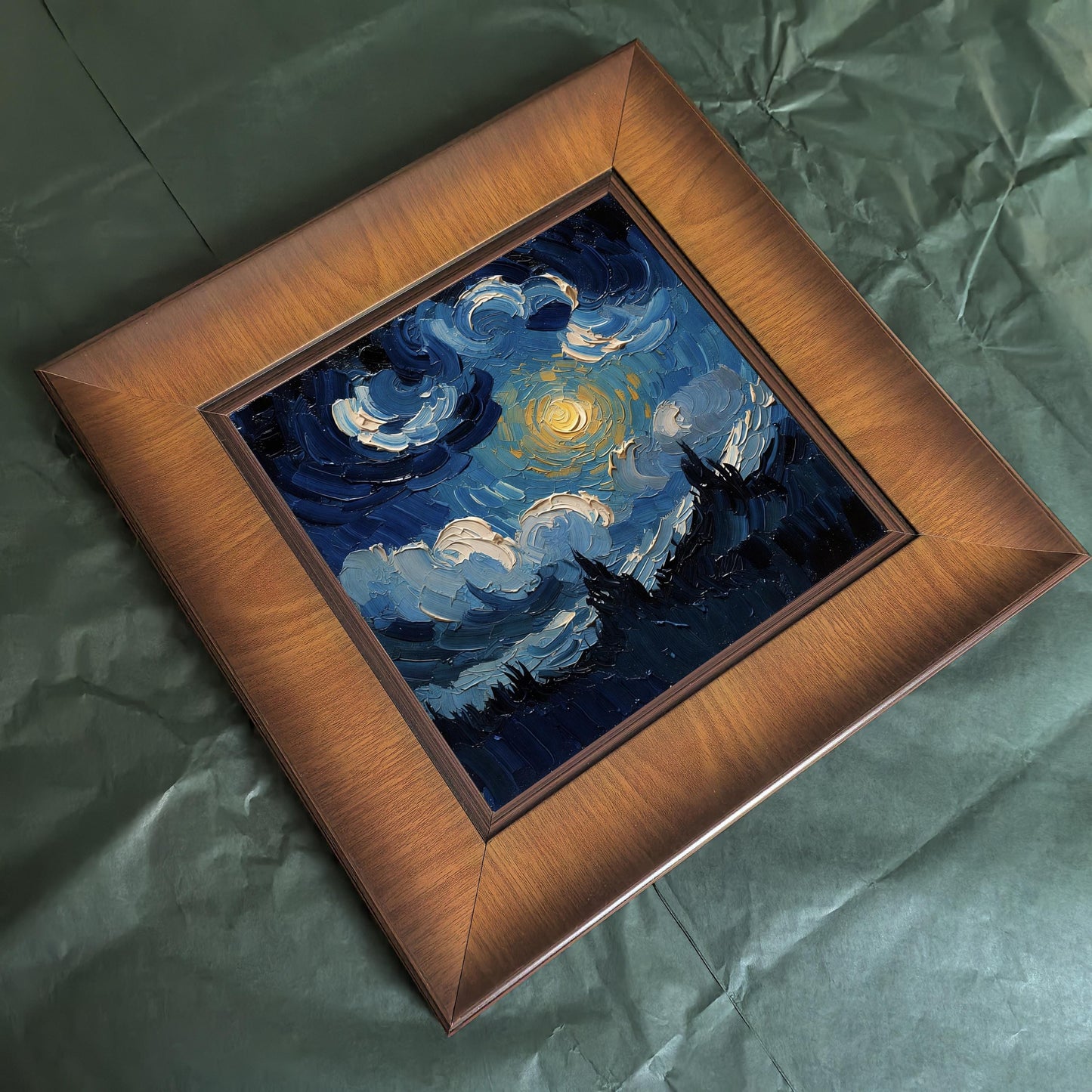 High-angle shot of a square framed textured painting with thick brushstrokes of a blue night sky and yellow moon, placed on a wrinkled green craft paper background.