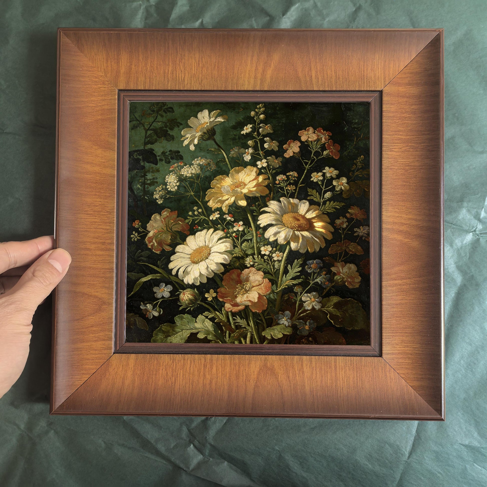 Close-up view of a hand holding a square wood-framed floral print, showcasing the rich grain of the wide wooden frame and the dark, moody background of the painting.