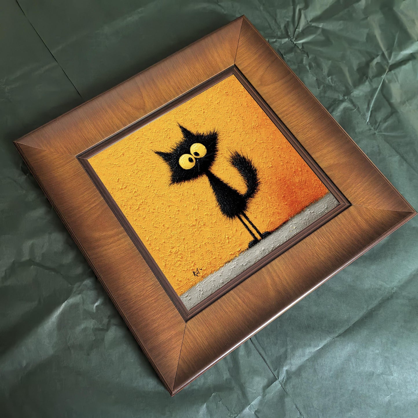 Overhead angled shot of a square wooden frame featuring a textured black cat painting, lying on a green paper background to highlight the wood grain.