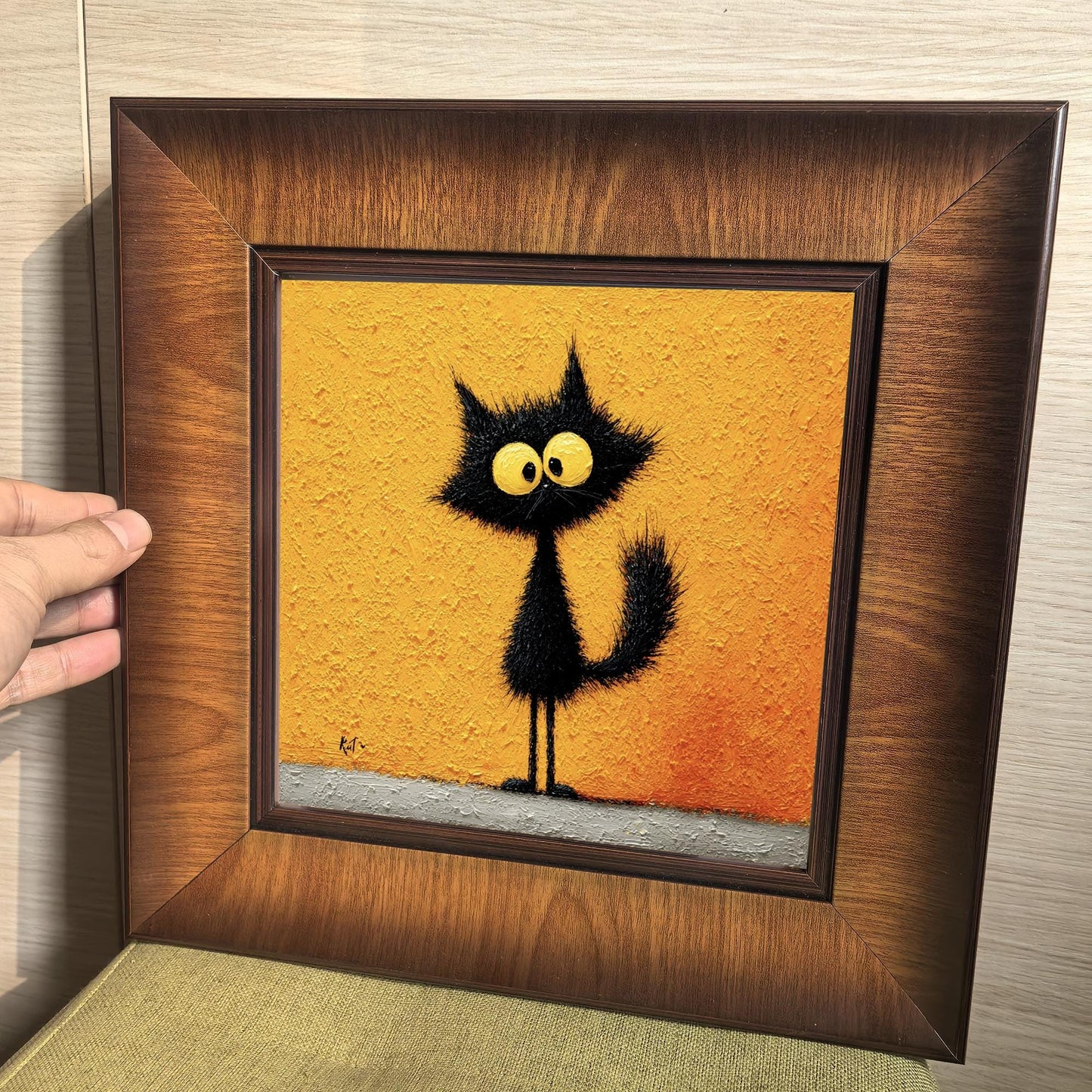 A hand holding a square wooden framed print of a whimsical black cat with large yellow eyes, showcasing the rich wood grain and the thick, impasto texture of the artwork.