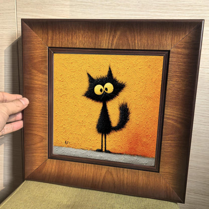 A hand holding a square wooden framed print of a whimsical black cat with large yellow eyes, showcasing the rich wood grain and the thick, impasto texture of the artwork.