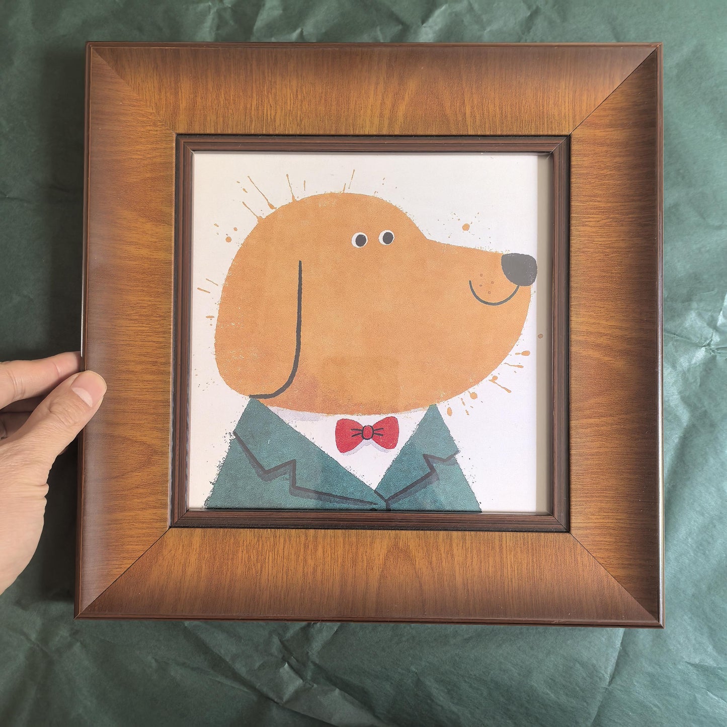 Close-up of a square wooden frame containing a minimalist yellow dog art print, held by a hand against a textured green background.