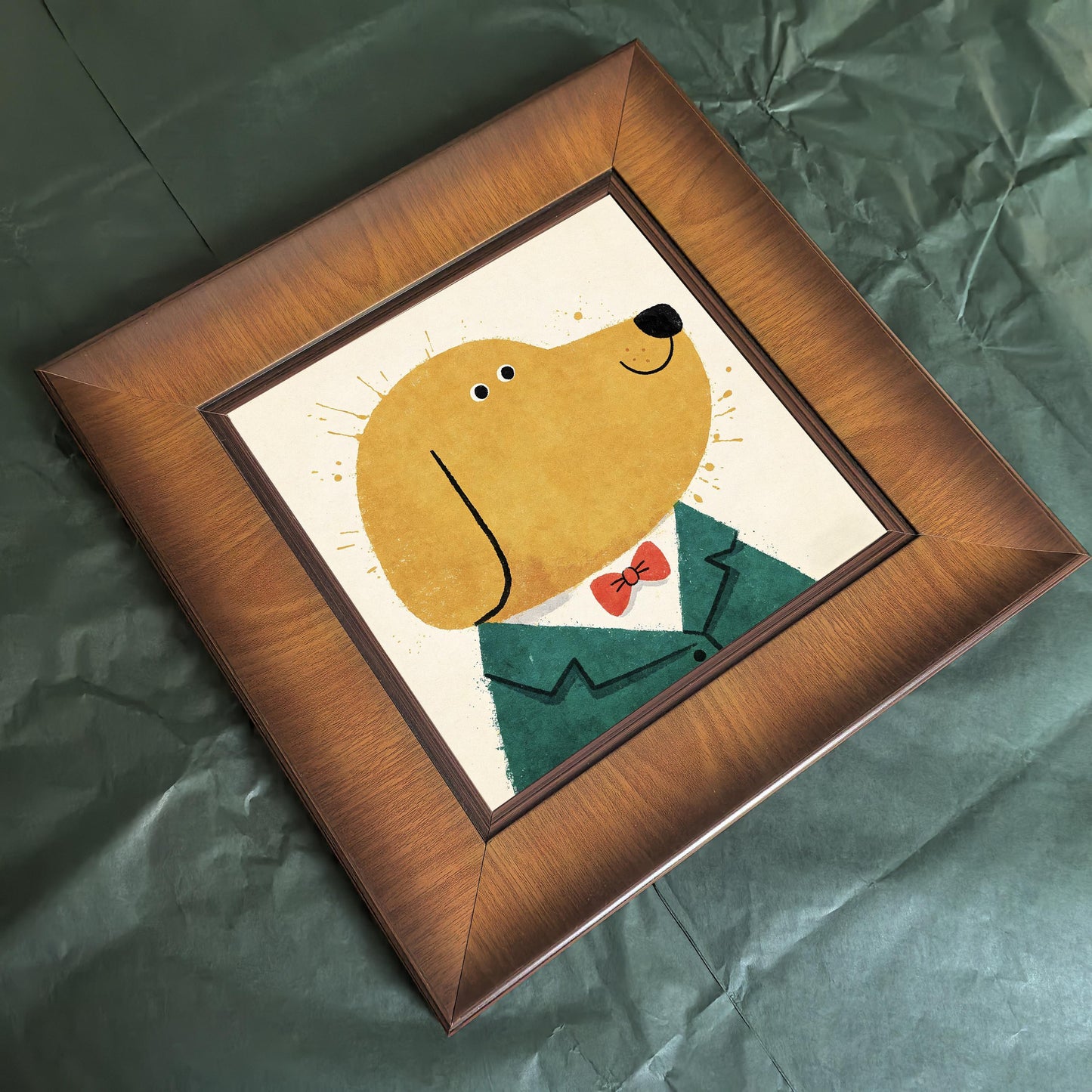 Overhead angled shot of a square wood-framed illustration of a dog wearing a red bowtie and green jacket, resting on crumpled green paper.