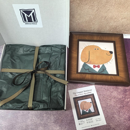 Premium unboxing experience for 'The Dapper Retriever' by Miracle Walls. A high-quality wood-framed print of a golden retriever in a suit and red bowtie sits beside a white gift box lined with green tissue paper and a gold-striped ribbon. An artist signature card detailing the museum-grade fine art paper and Giclée printing is displayed in the foreground.
