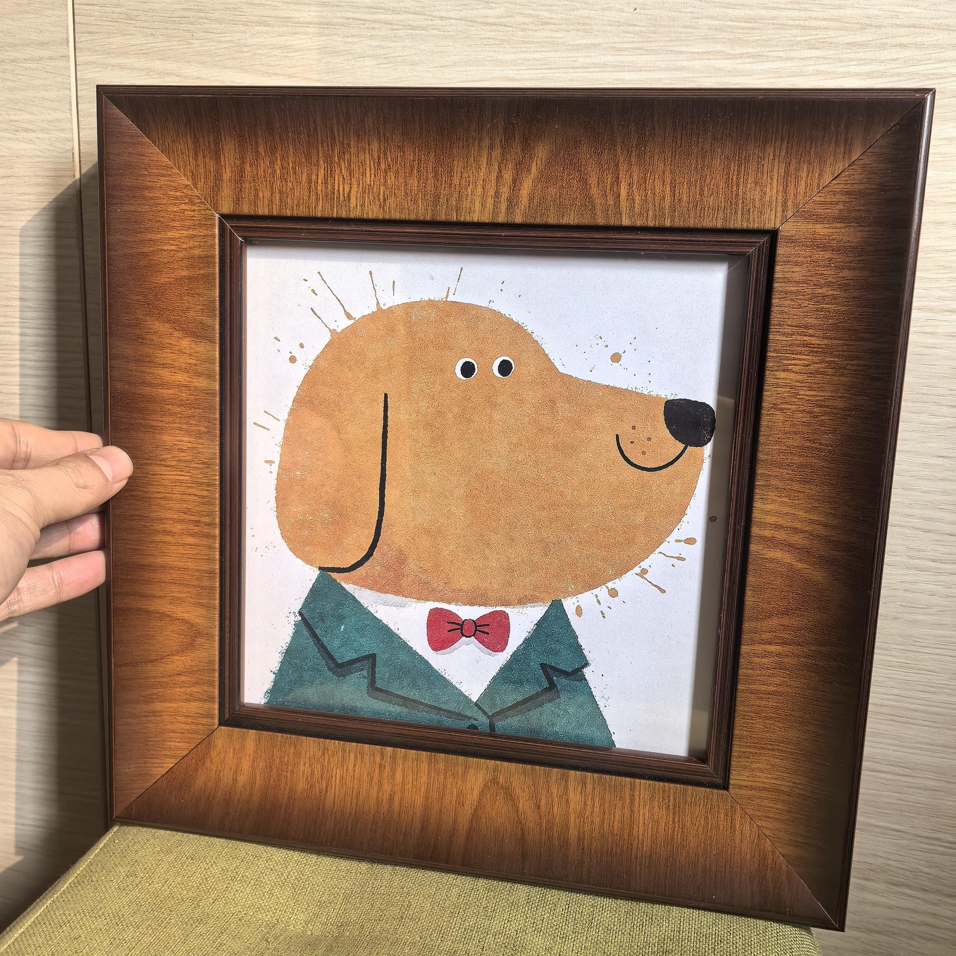 A hand holding a square wood-framed art print of a dog in a suit, showing the rich grain of the wide wooden frame against a light wood wall.