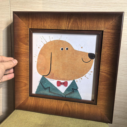 A hand holding a square wood-framed art print of a dog in a suit, showing the rich grain of the wide wooden frame against a light wood wall.