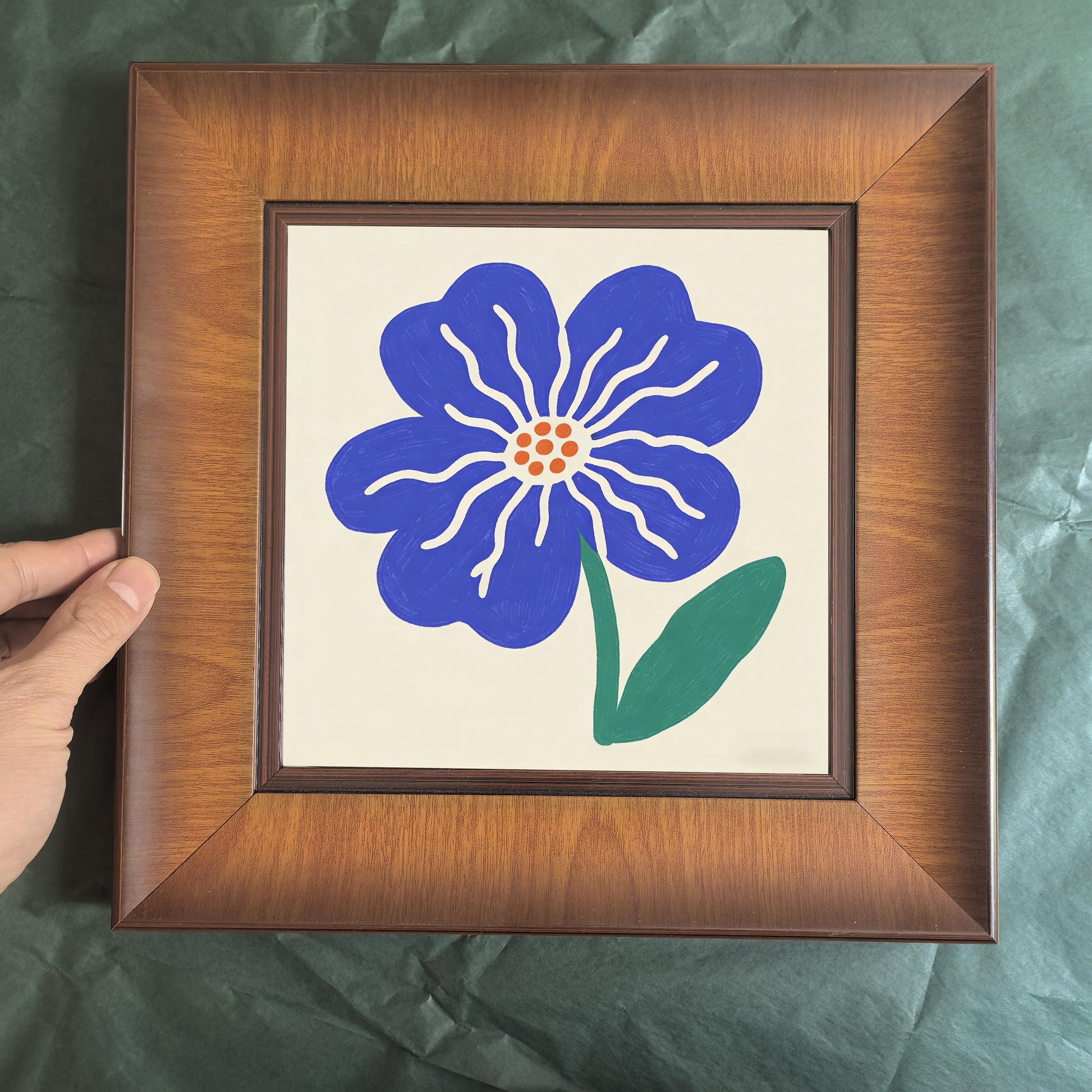 Front-facing shot of a square wooden picture frame featuring a blue hand-painted style flower, held by a person against a green background.