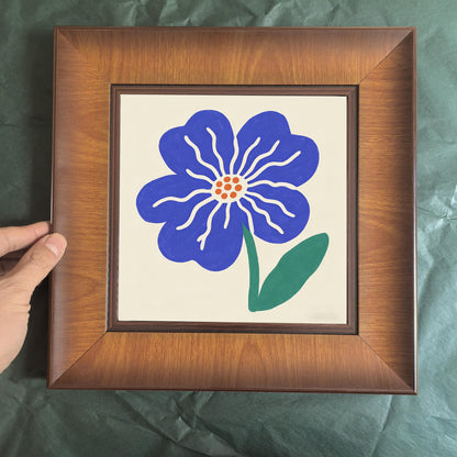Front-facing shot of a square wooden picture frame featuring a blue hand-painted style flower, held by a person against a green background.